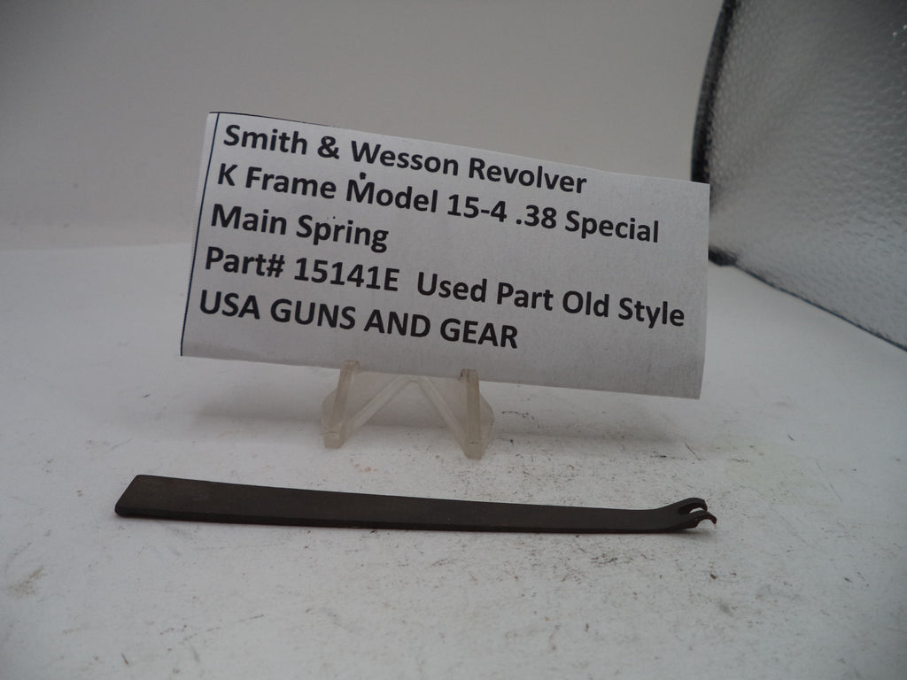 A used metal main spring part for a Smith & Wesson revolver, displayed on a small clear plastic stand with a white paper label showing the part number and description.