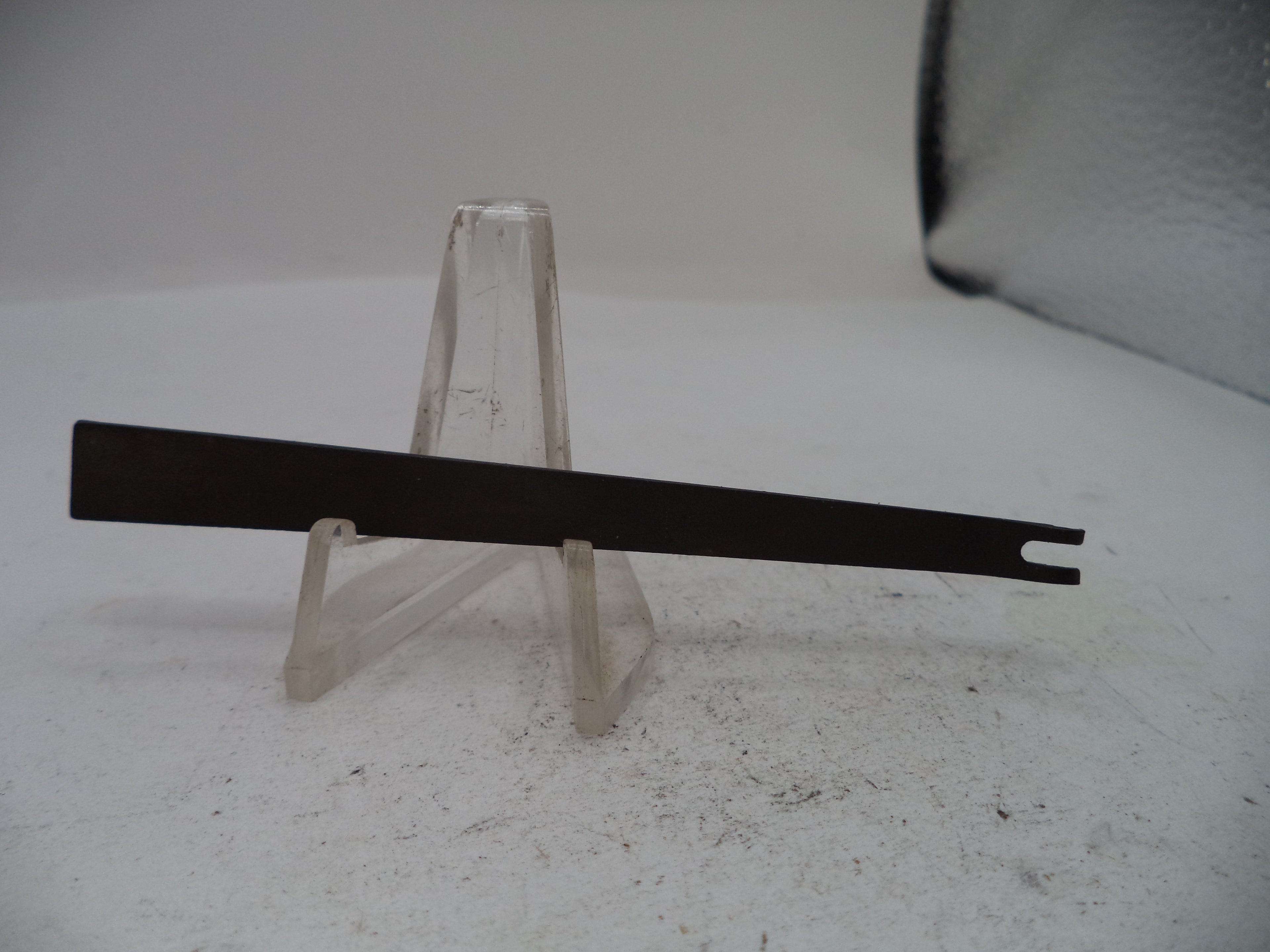 A flat metal tool with a single hook-like end, displayed horizontally on a clear plastic stand against a light gray background.