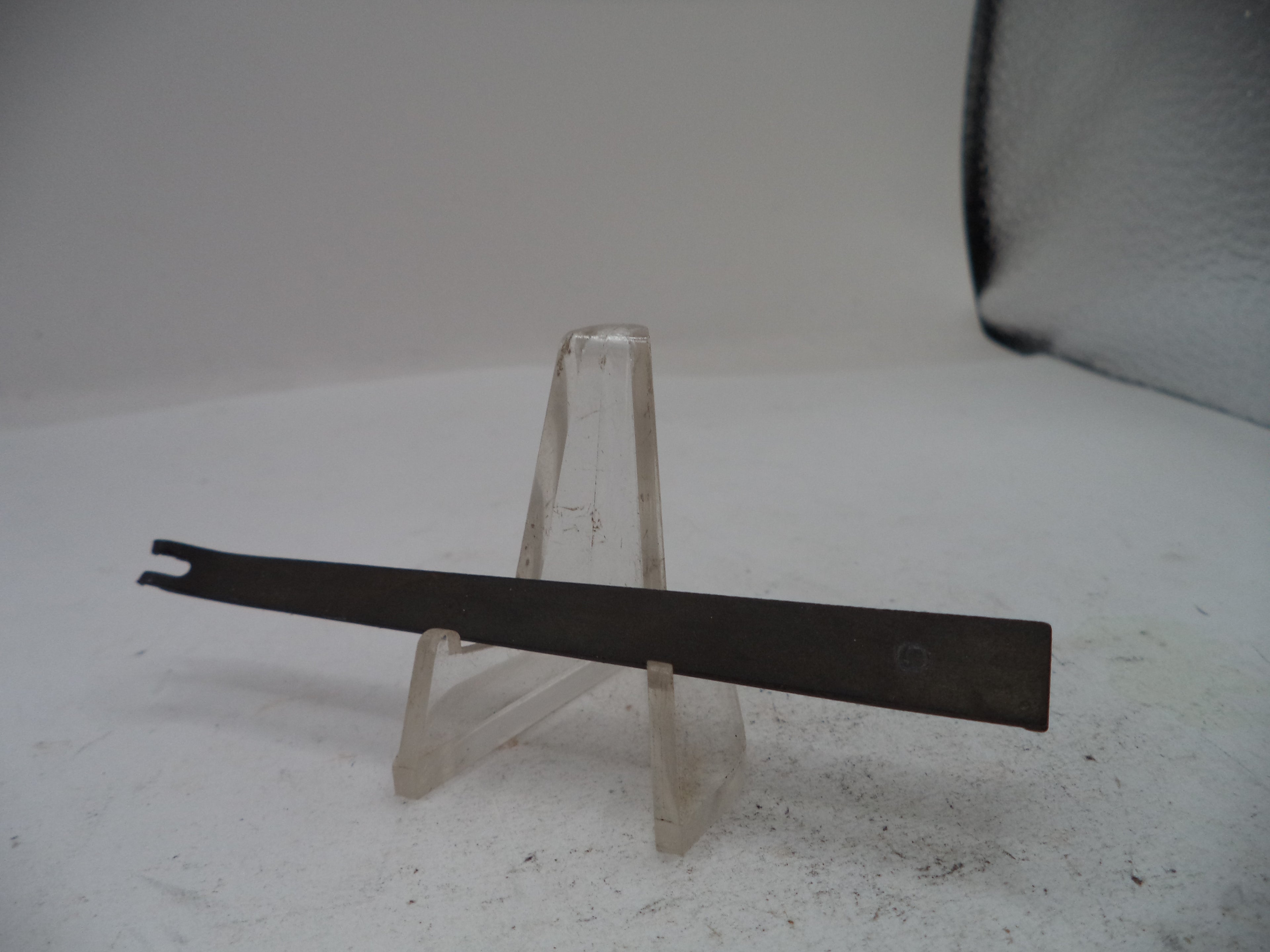 A flat metal tool with a forked tip, displayed horizontally on a clear plastic stand against a plain light background.