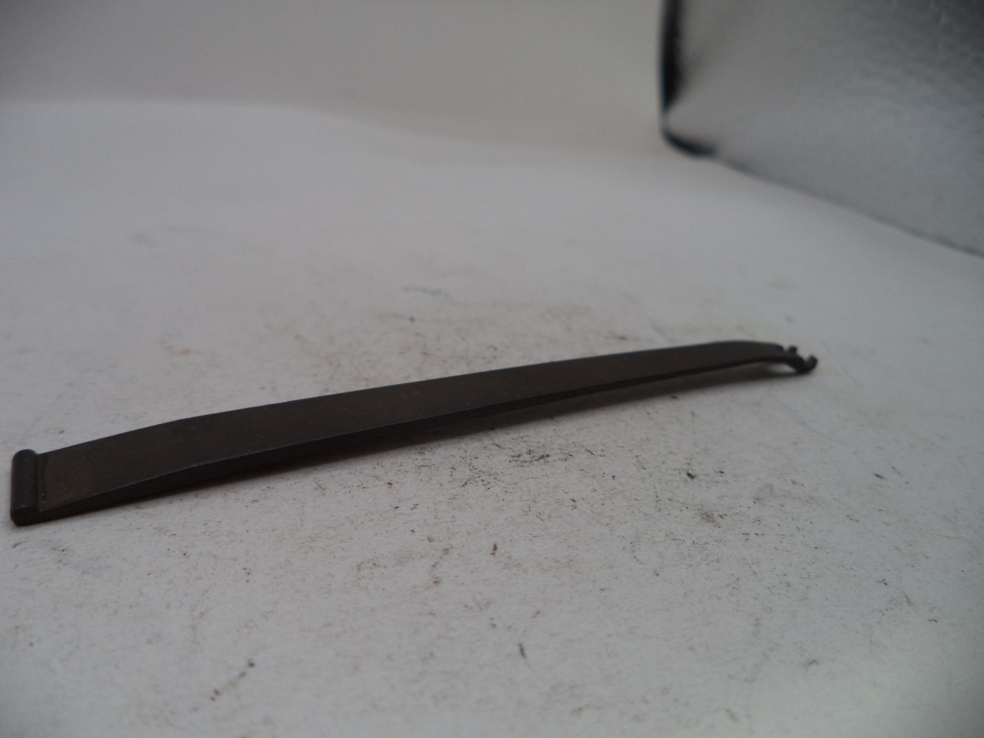 A long, thin black metal tool with a flat blade and a small hook-like end, photographed on a light gray surface.