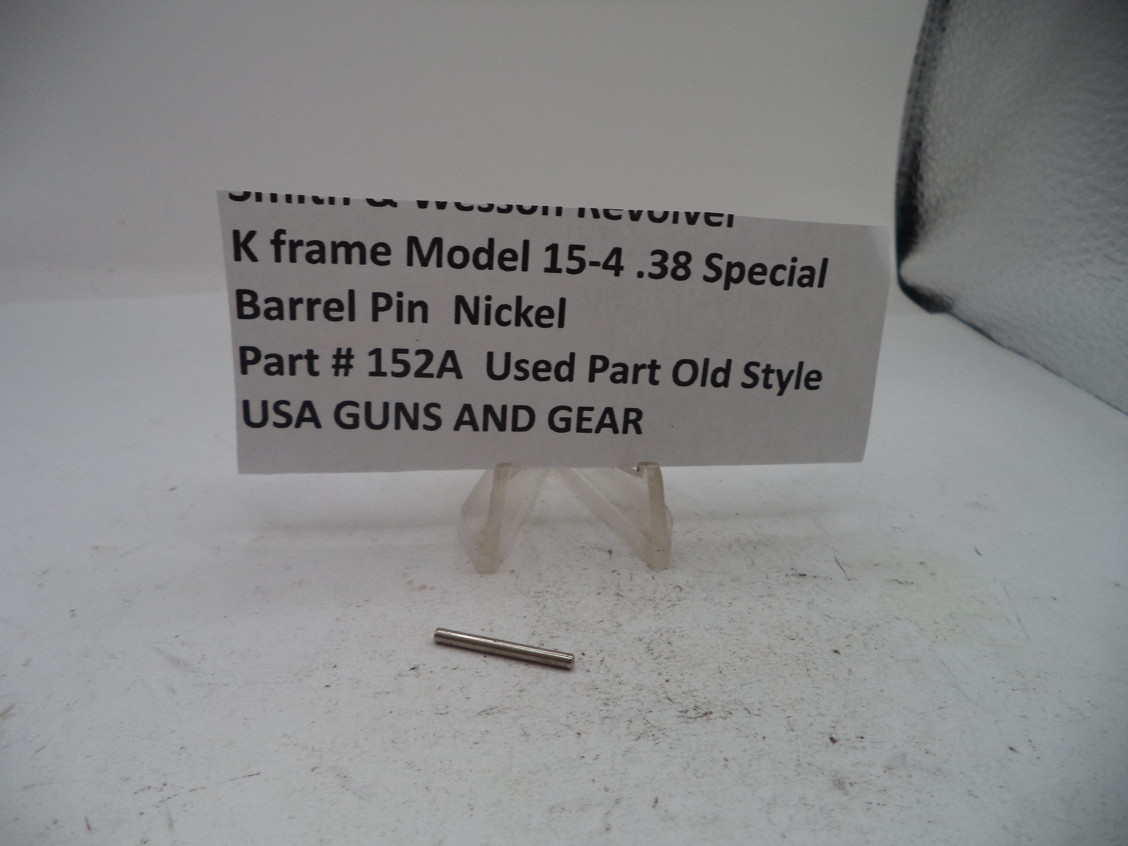 A small metallic nickel barrel pin displayed on a white surface next to a paper label that reads 'Smith & Wesson Revolver K frame Model 15-4 .38 Special Barrel Pin Nickel Part # 152A Used Part Old Style USA GUNS AND GEAR'.