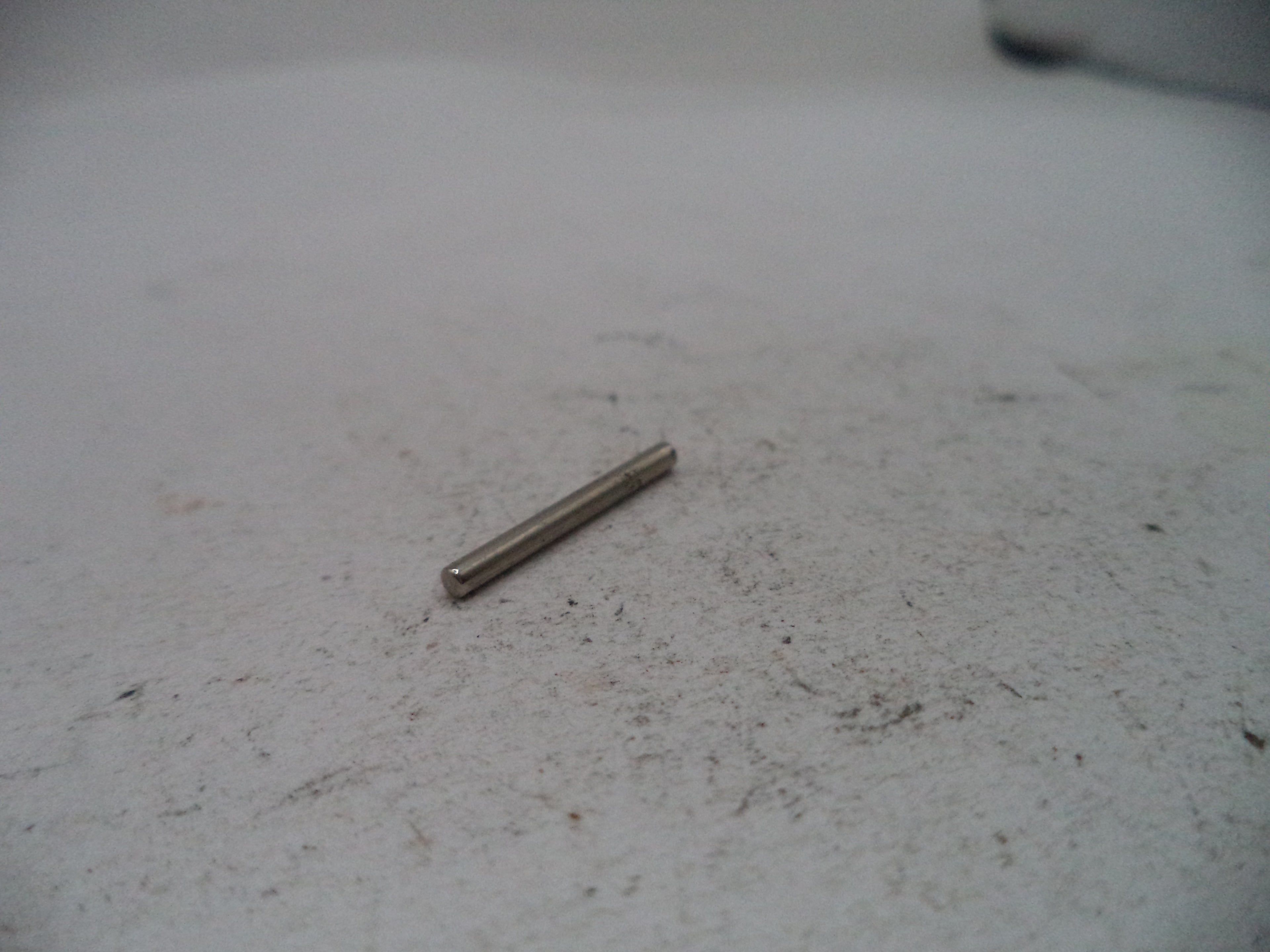 A small metallic cylindrical pin or rod lying on a light gray textured surface.