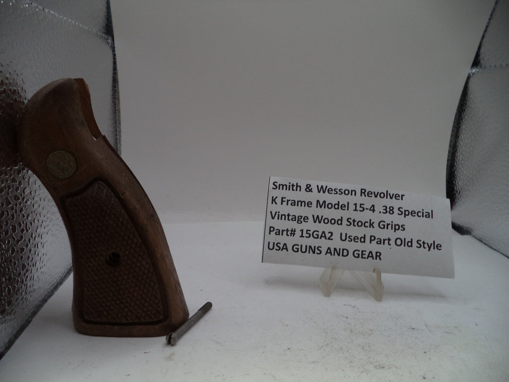 A vintage Smith & Wesson revolver K-frame stock grip part shown standing upright next to a small white label card on a white surface, with a textured silver backdrop.