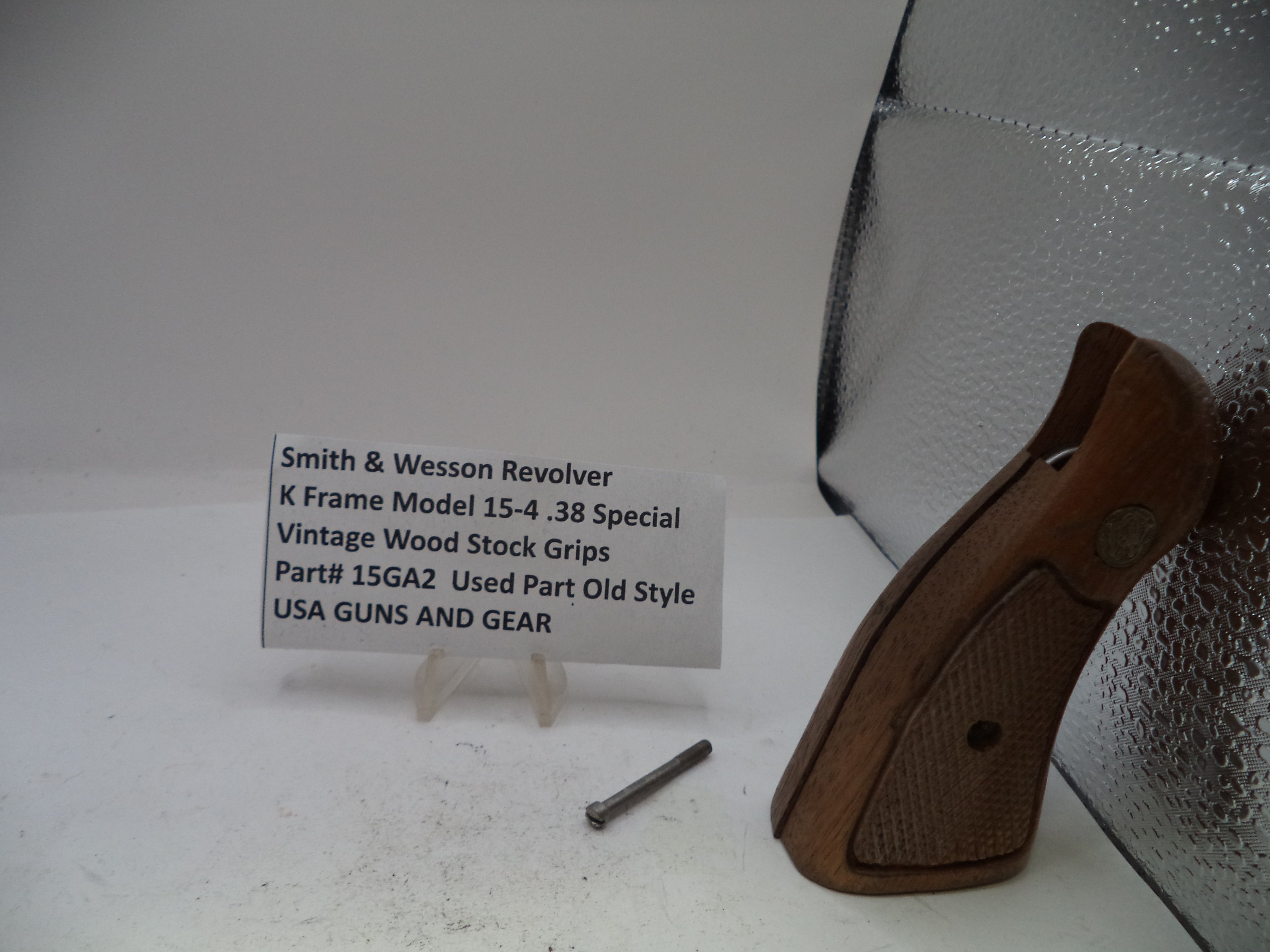 A used vintage wood stock grip for a Smith & Wesson revolver, displayed next to its metal mounting screw on a white surface with a small label card.