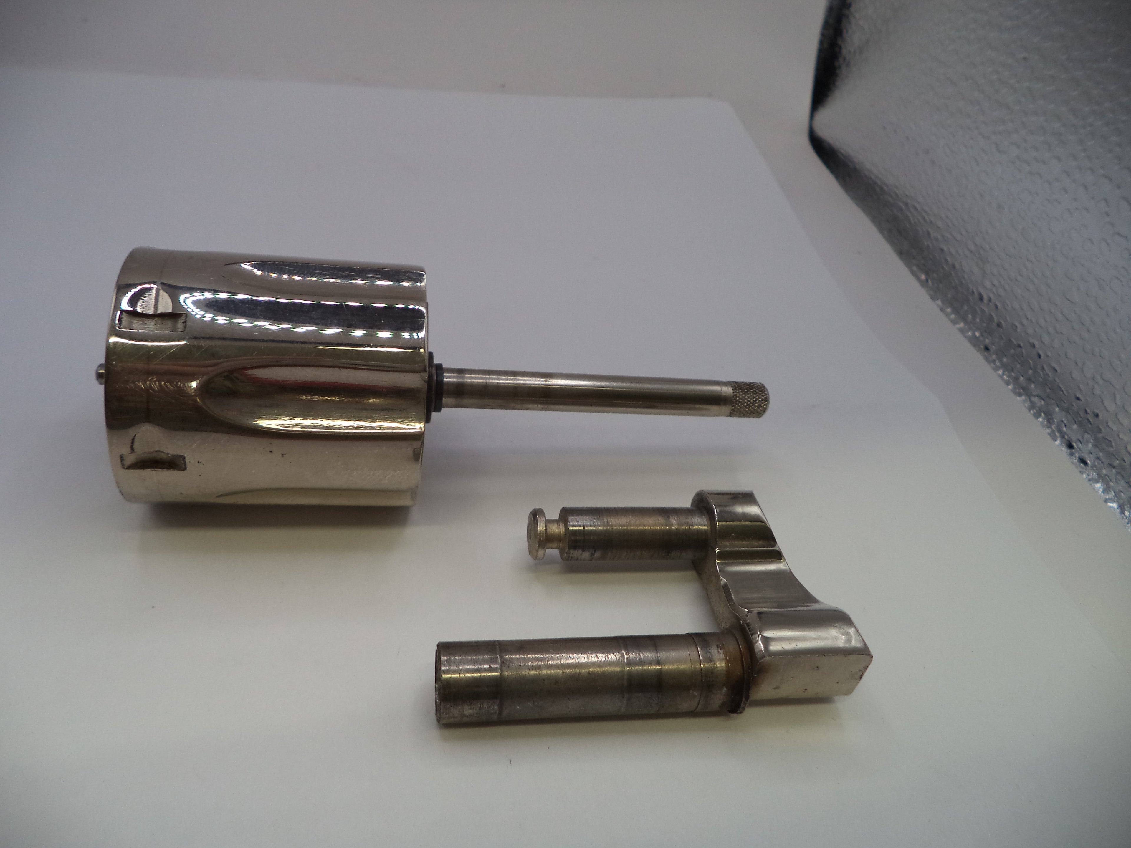 A polished metallic cylindrical component with a long horizontal rod, shown alongside a separate matching metal bracket on a plain white surface.