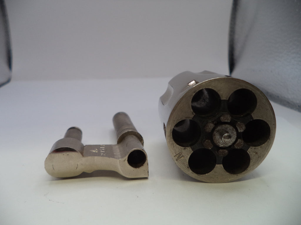 A close-up of a metallic revolver cylinder disassembled to show the six internal chambers, alongside a matching metal component on a plain light background.