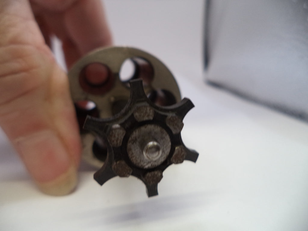 A close-up of a hand holding a small metal mechanical component with a dark star-shaped gear and a circular metal disc against a white background.