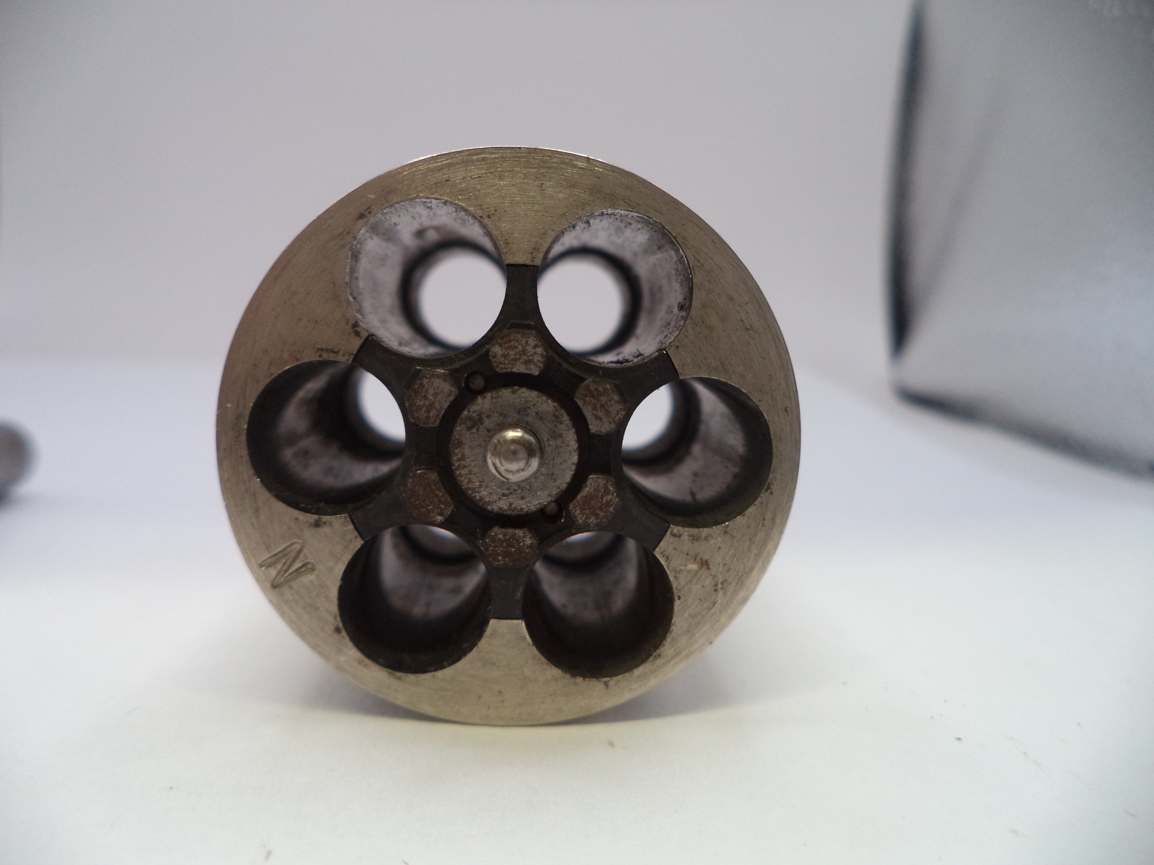 A close-up of a metallic revolver cylinder base with six chambers visible, showing a weathered silver and dark gray finish on a white surface.