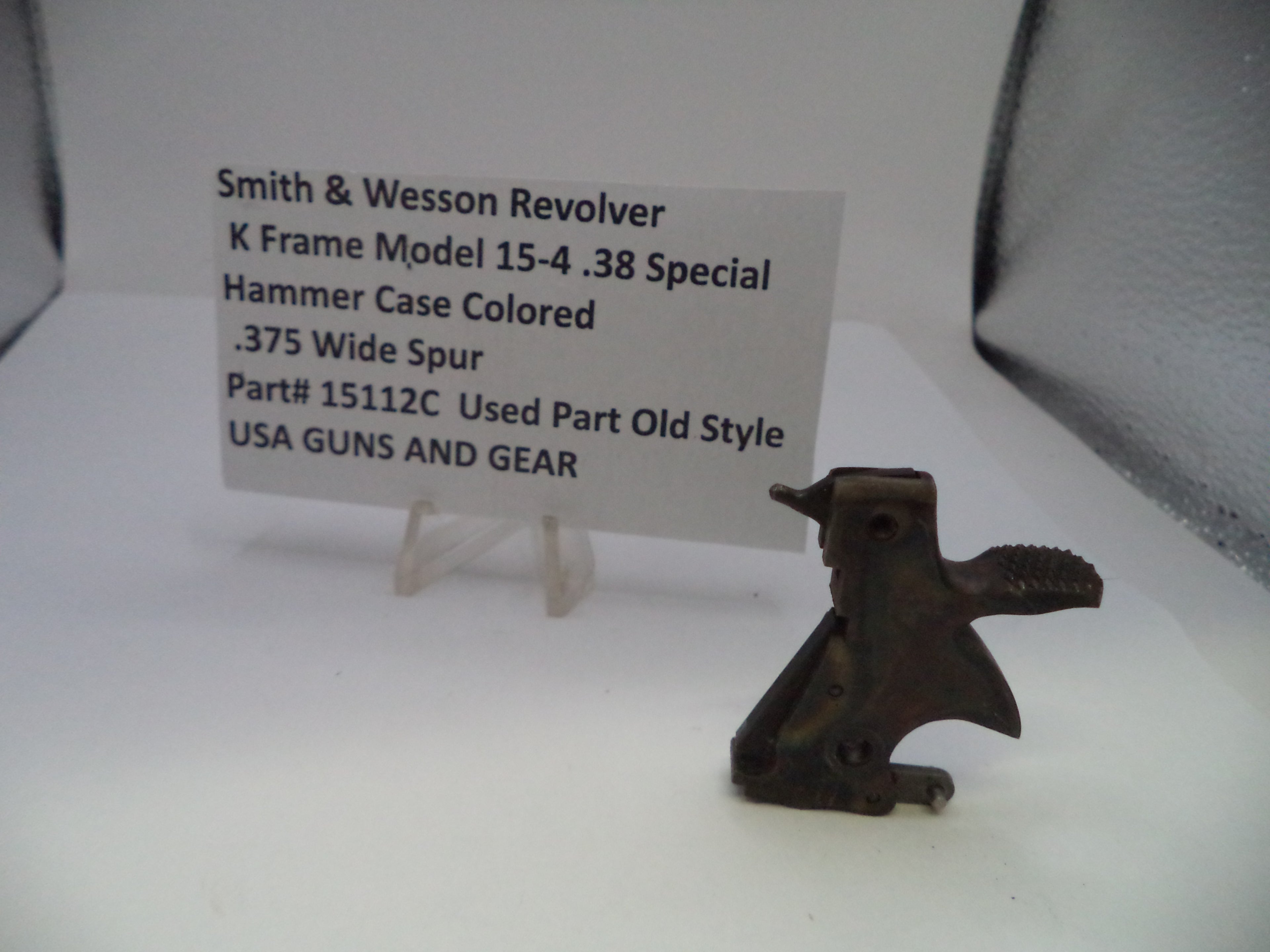 A used metal hammer case part for a Smith & Wesson revolver, displayed next to a white label card on a clear stand against a white background.