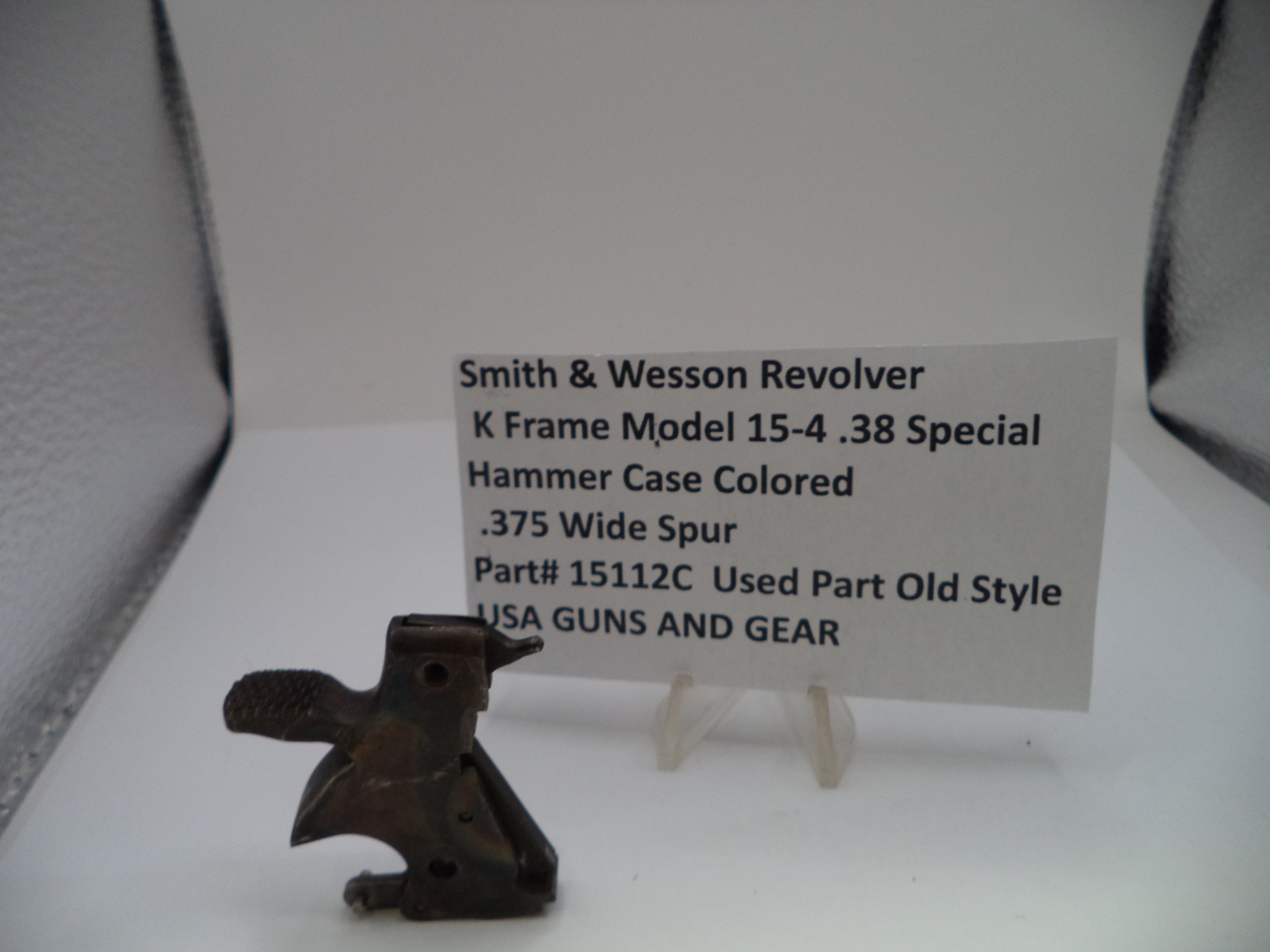 A small dark metal revolver hammer case part displayed on a white surface with a white label card behind it reading 'Smith & Wesson Revolver K Frame Model 15-4 .38 Special Hammer Case Colored .