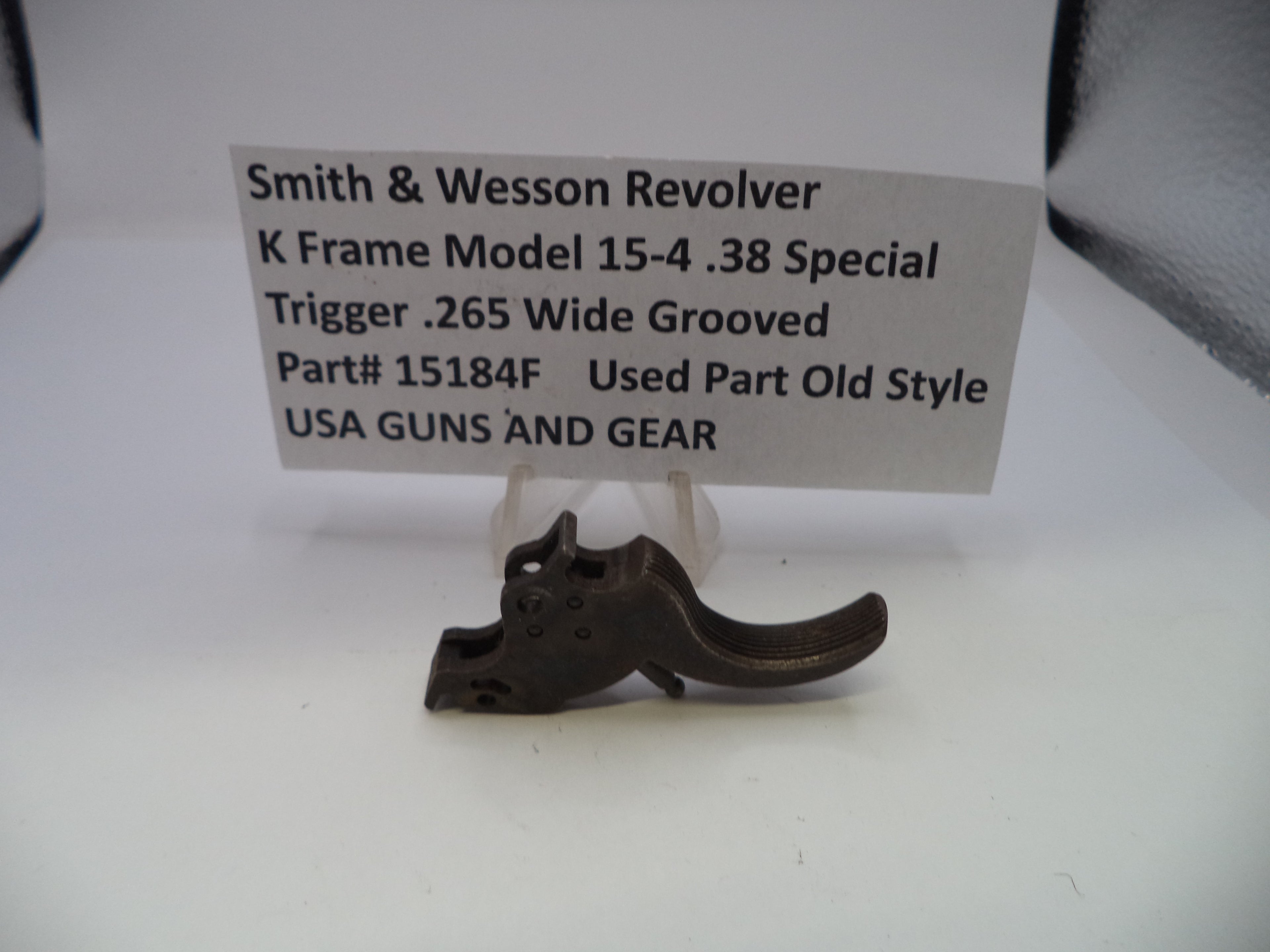 A small dark metal Smith & Wesson revolver trigger part displayed on a clear stand in front of a white label card with printed product details.
