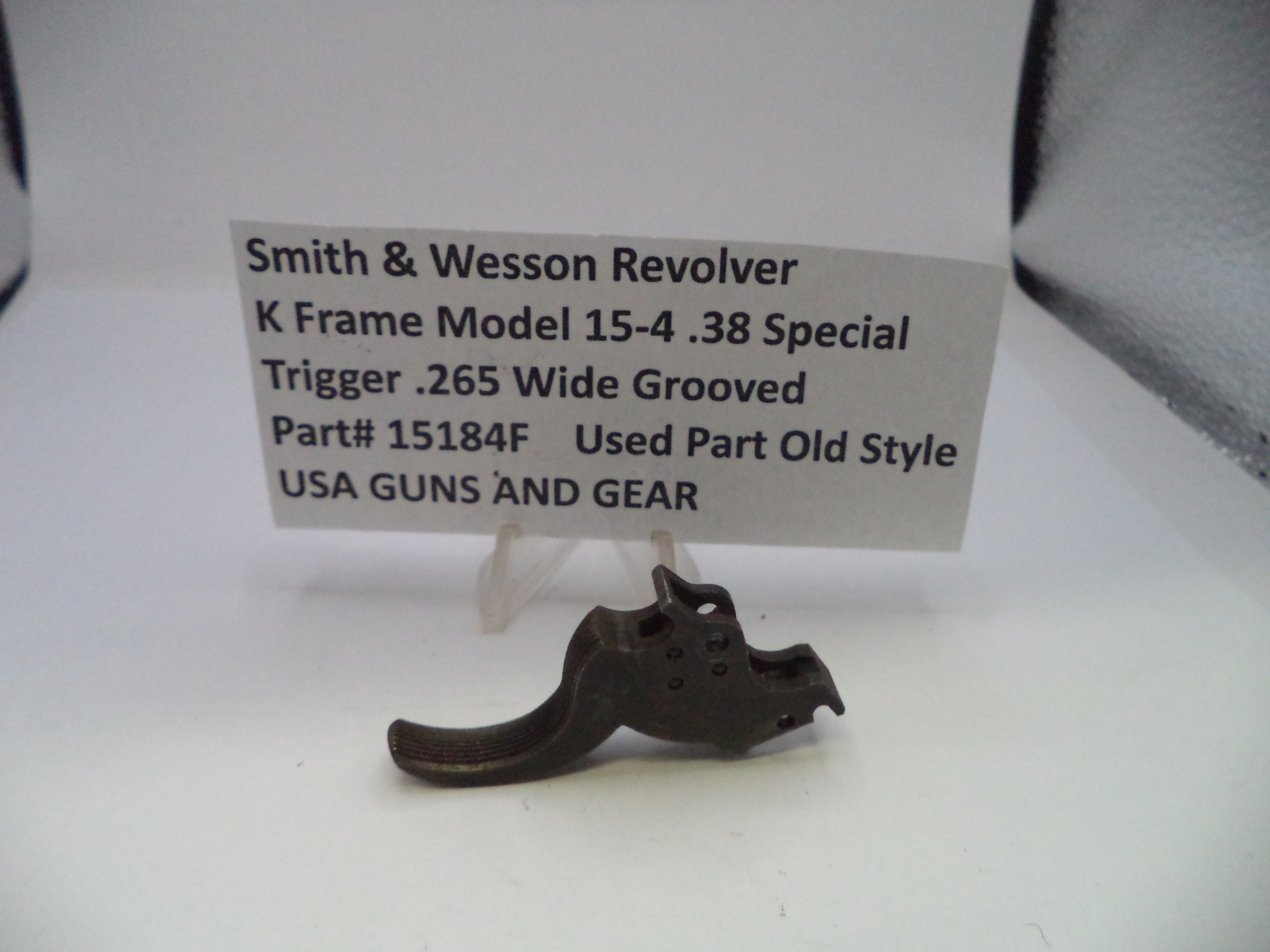 A used metal trigger component for a Smith & Wesson revolver, displayed on a white surface with a paper label card behind it.