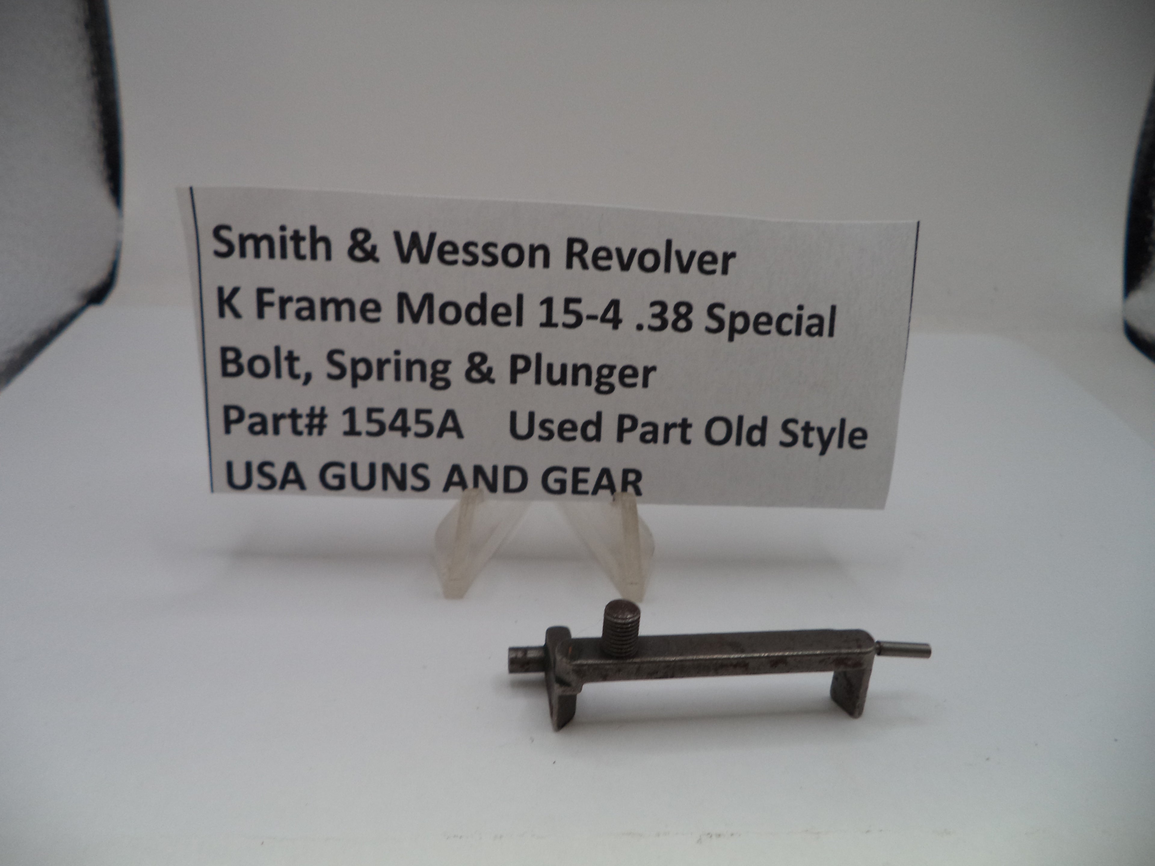 A used metal Smith & Wesson revolver bolt, spring, and plunger part, displayed on a small clear stand in front of a white paper label card with product details.