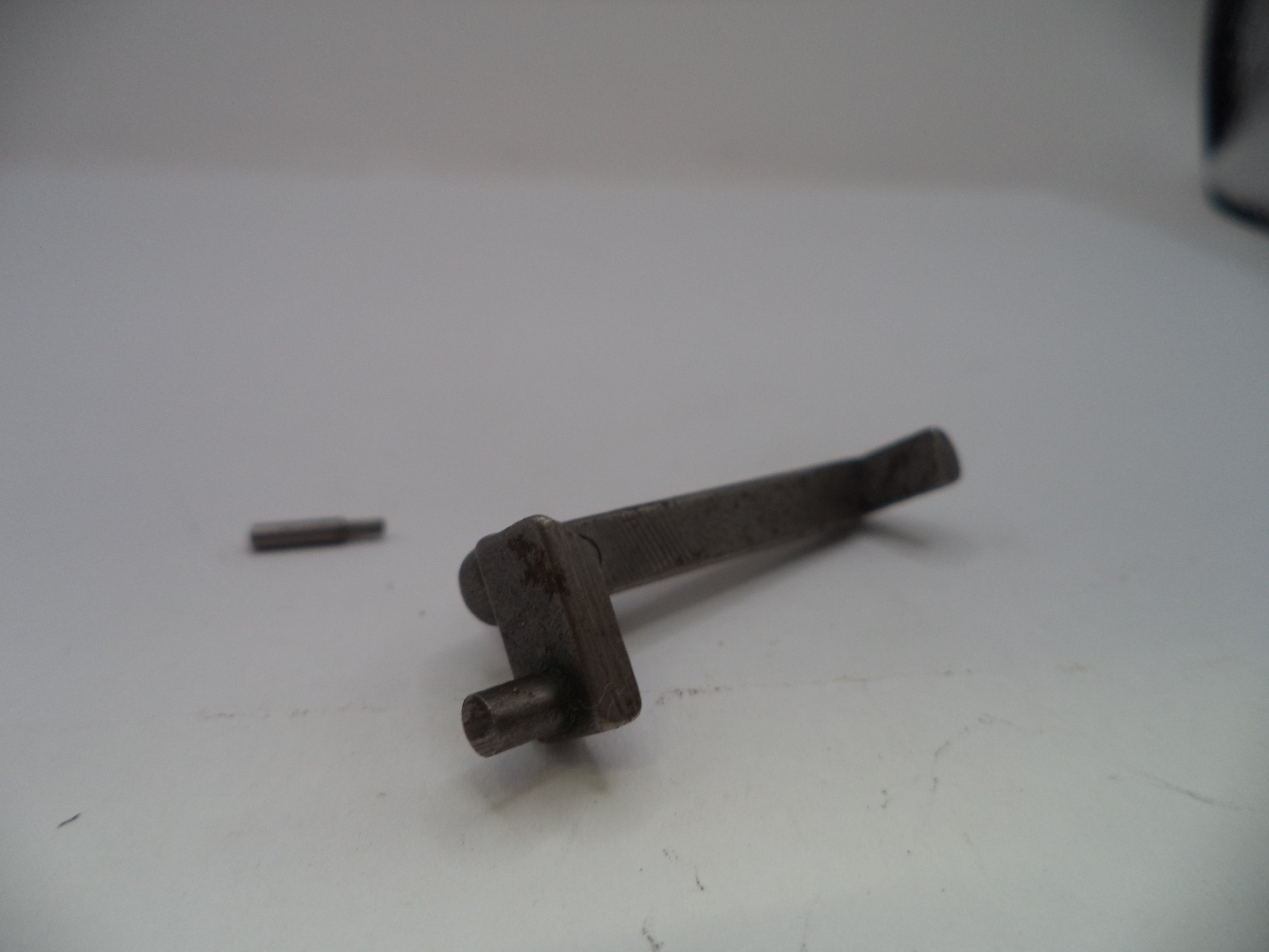 A small dark metal mechanical part or tool component with a short cylindrical pin and a bent stem, photographed on a plain light background with a separate small pin nearby.