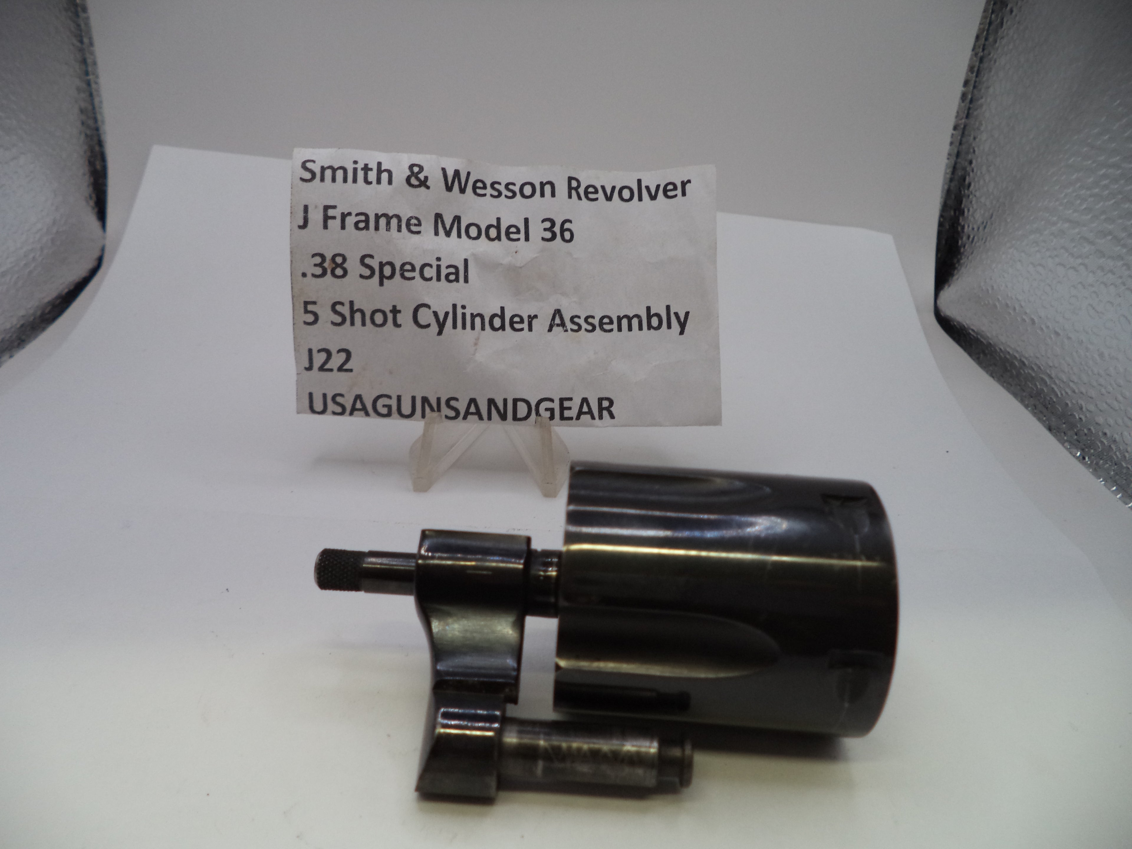 A black metal Smith & Wesson revolver cylinder assembly with a small clear plastic display stand and a paper label card behind it, photographed on a white surface with a gray backdrop.