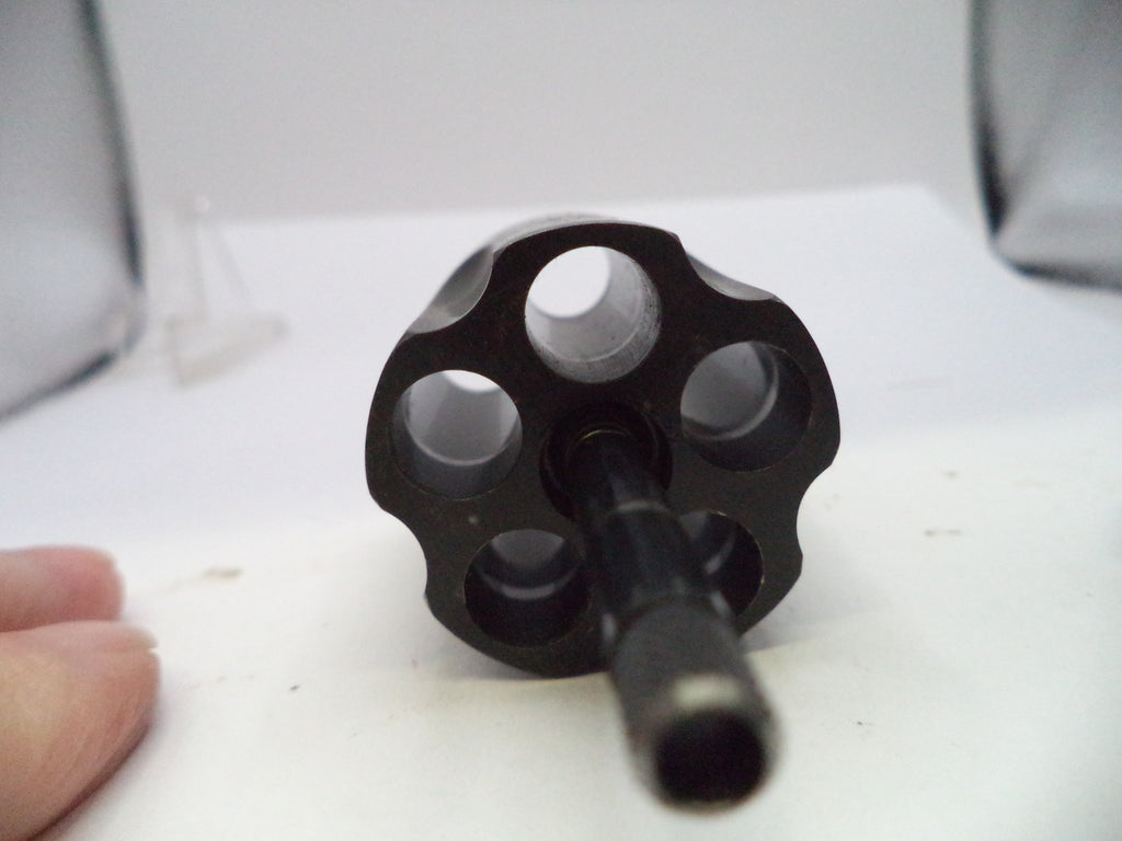 A black metal six-hole revolver cylinder or barrel component shown close-up on a white surface, with a finger partially visible at the lower left for scale.