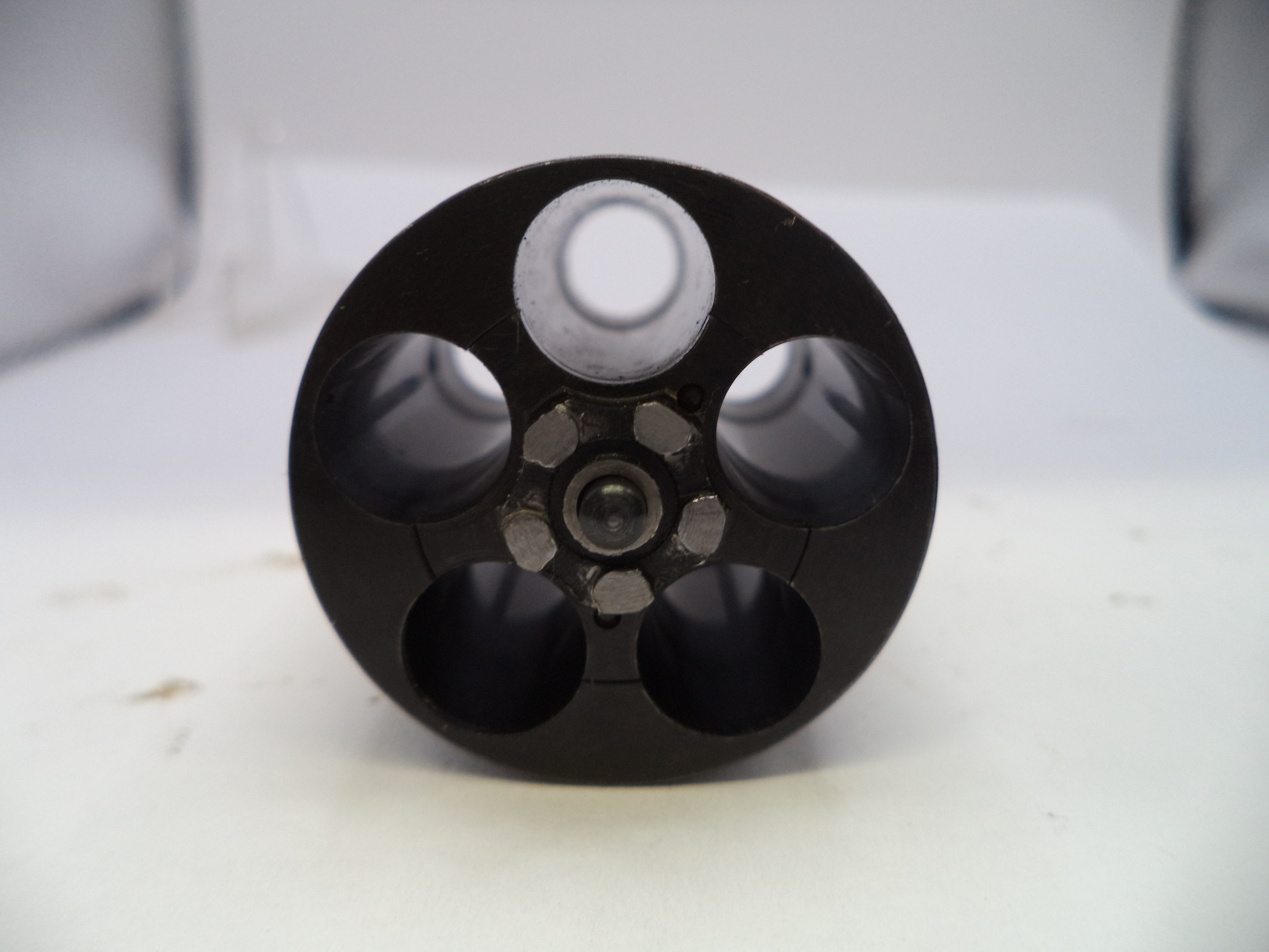 A close-up of a black circular metal component with five circular openings, photographed on a white surface with a soft-focus light gray background.