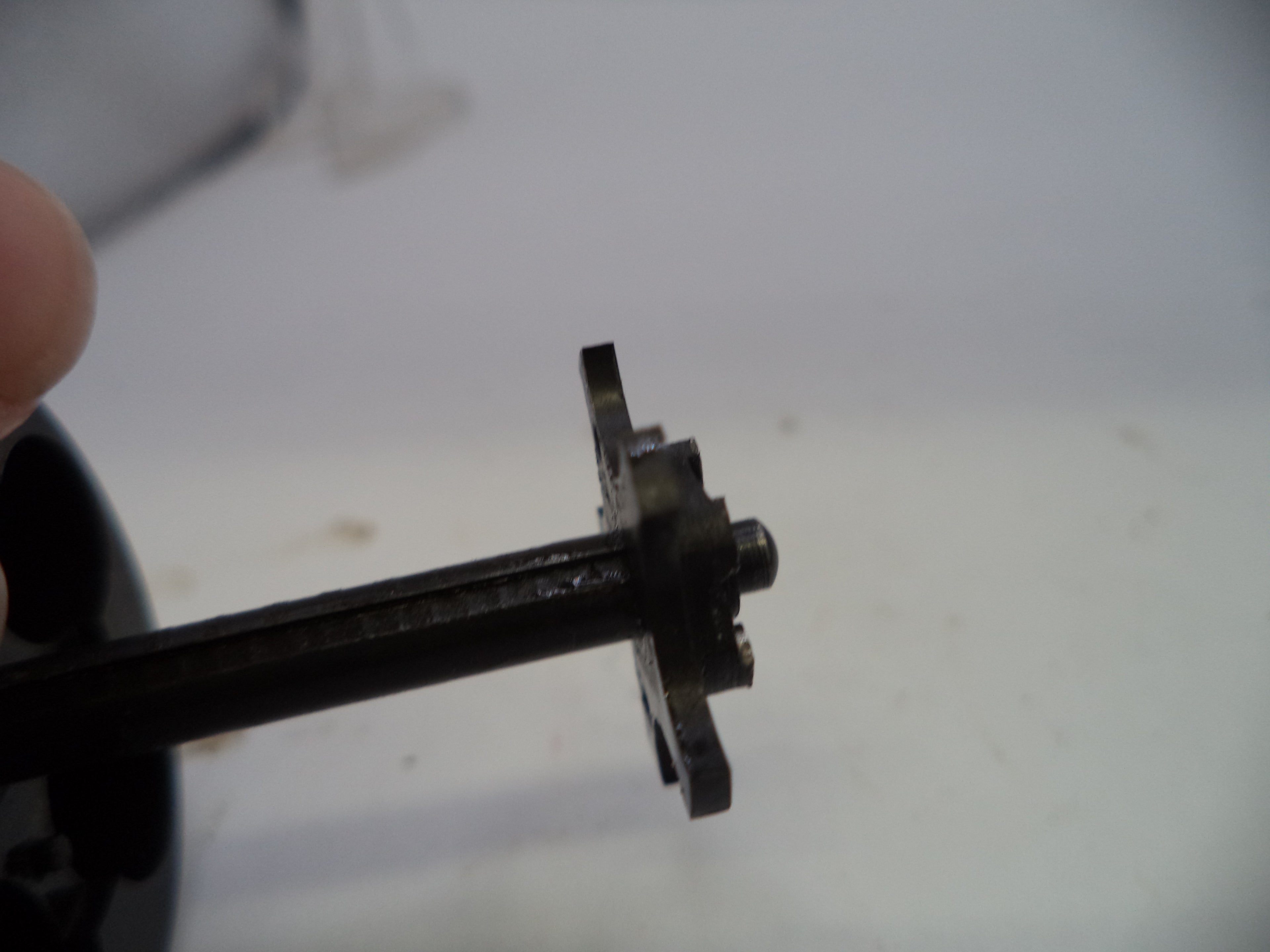 A close-up of a small black metal mechanical component or gear assembly held by a finger against a plain light background.
