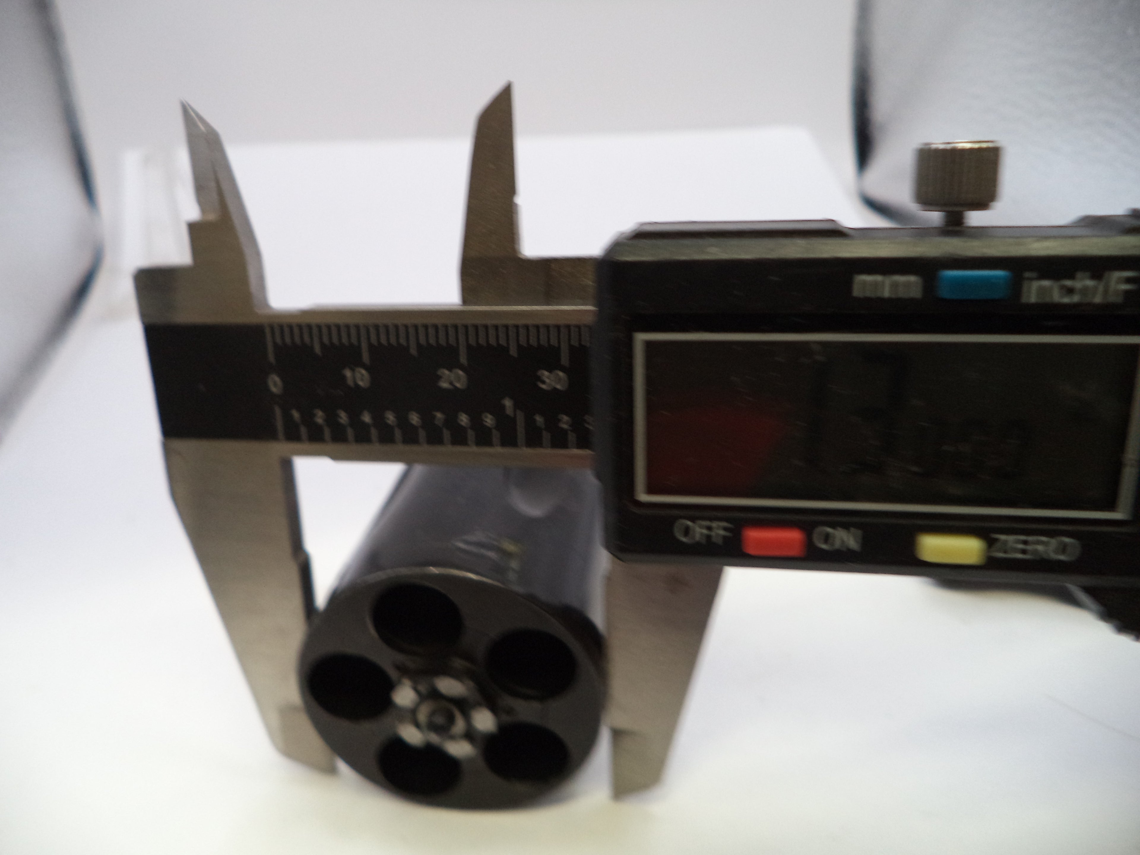 A digital caliper measuring the diameter of a black cylindrical object with four circular holes, shown on a white surface.