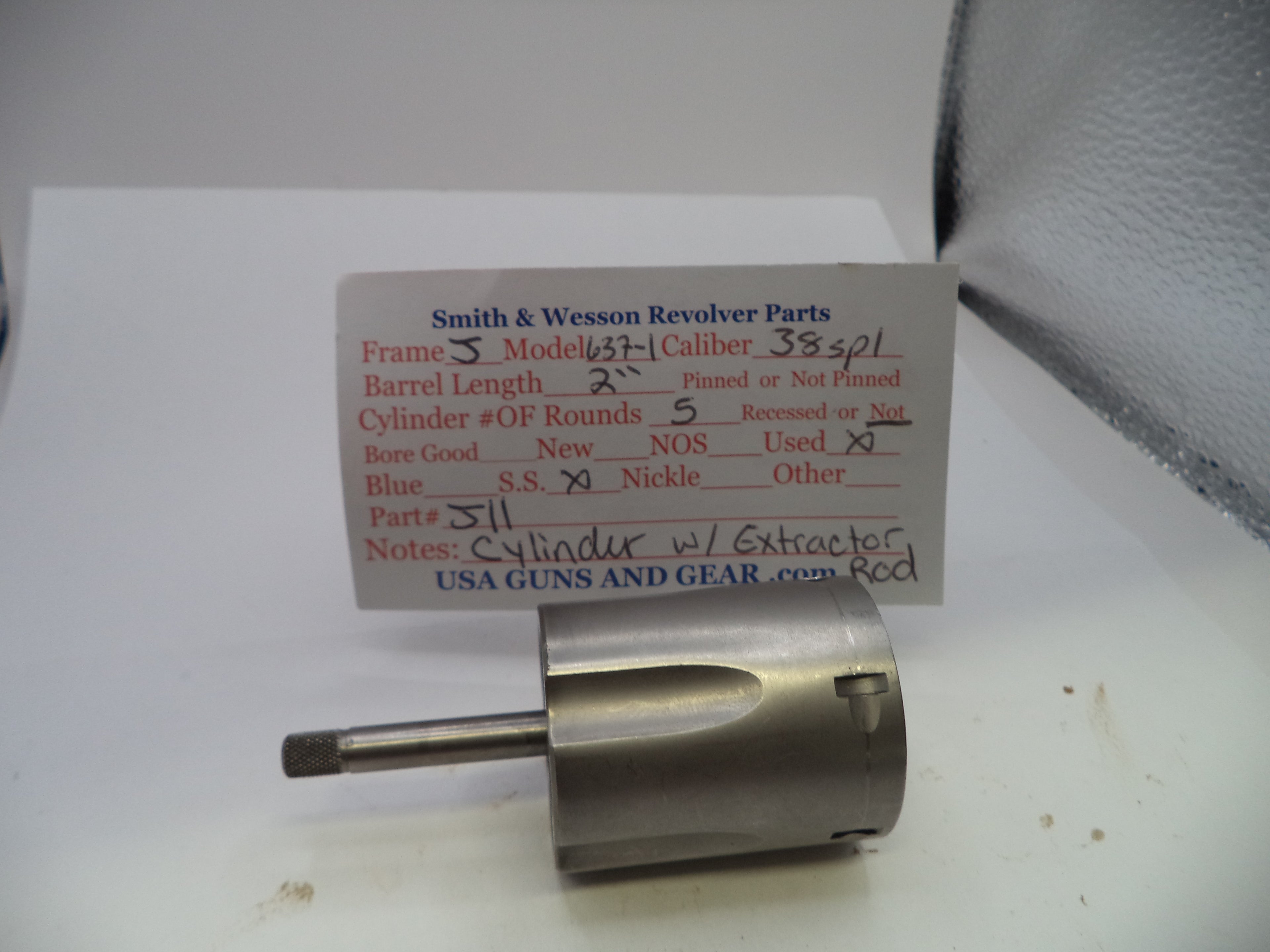 A used metal revolver cylinder and extractor assembly from Smith & Wesson, shown with its original handwritten parts identification card on a white surface.
