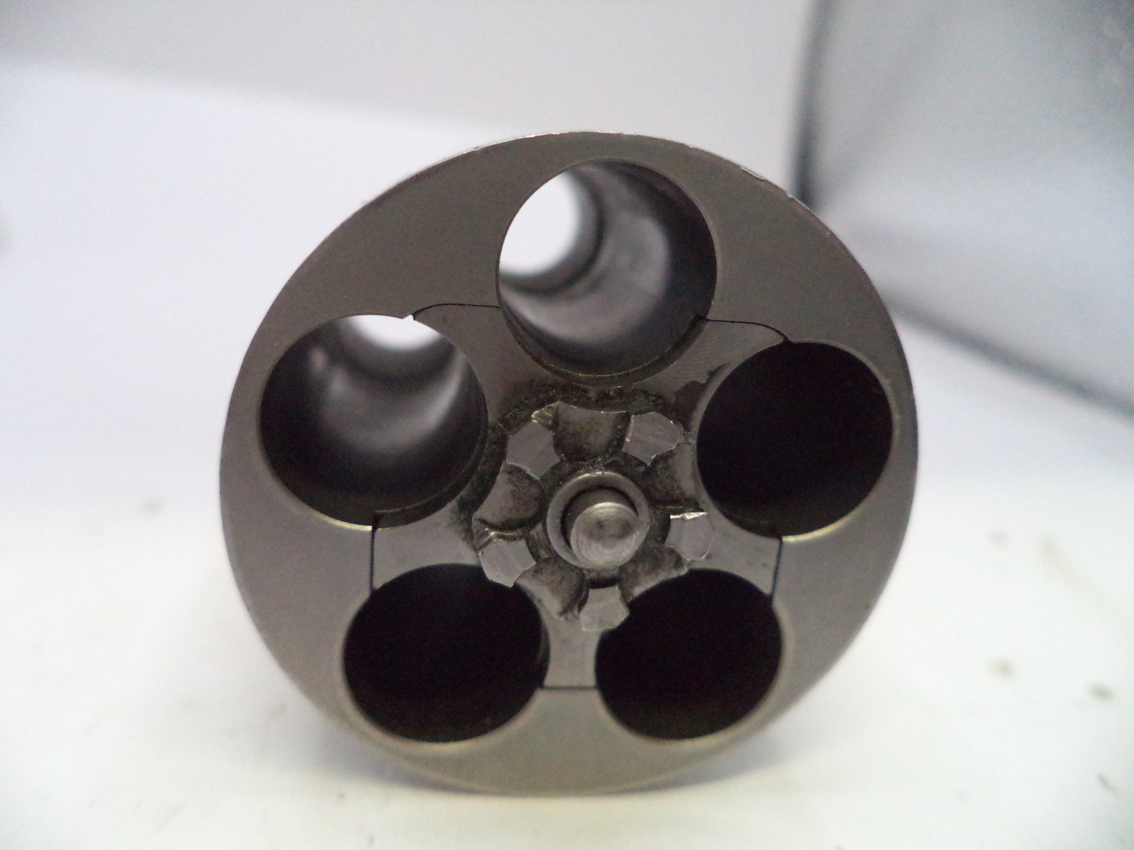 A close-up of a metallic circular component with four large dark openings and a central threaded or textured hub, photographed on a light gray to white background.