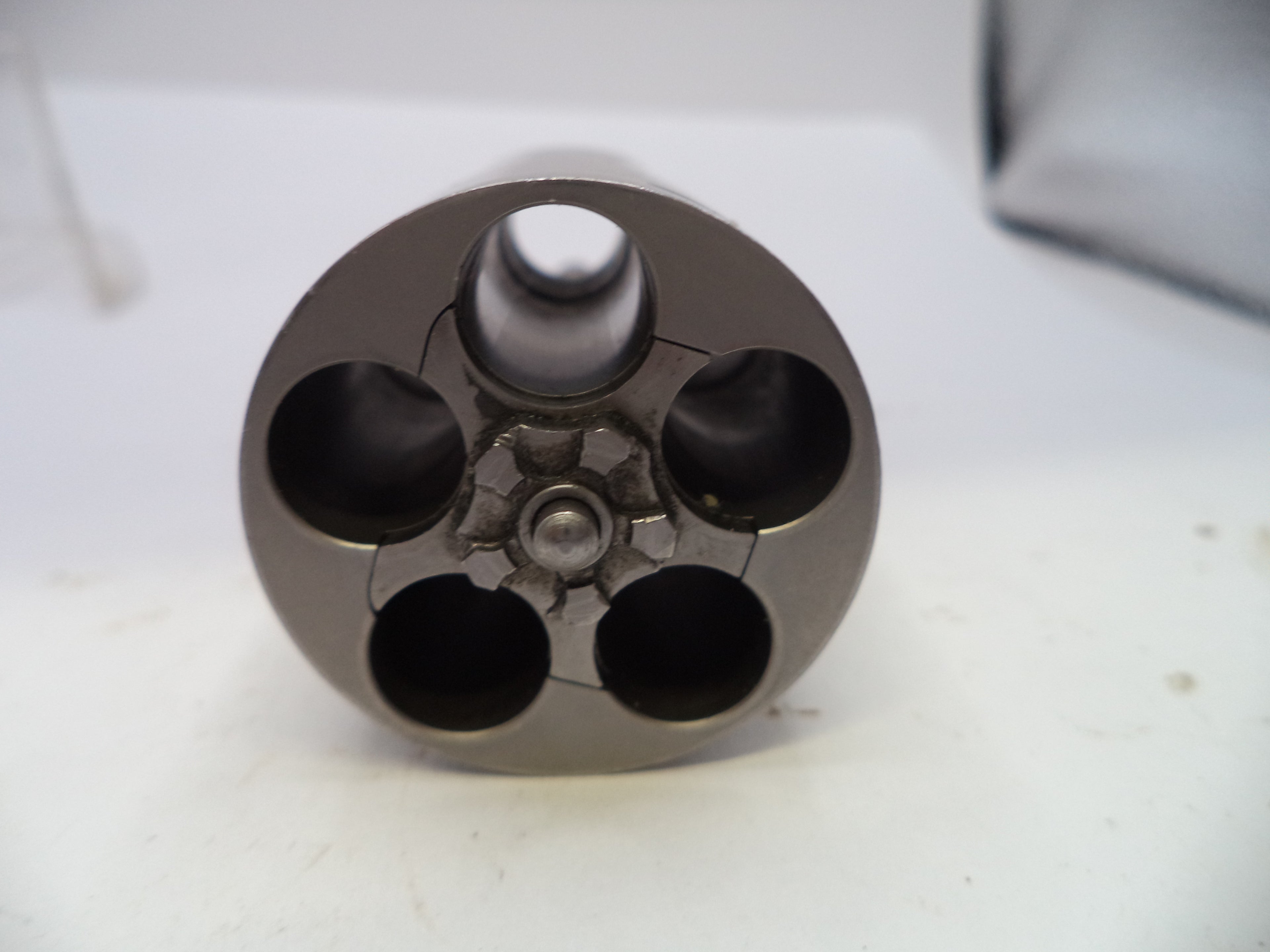 A close-up of a metallic cylindrical component with four circular openings, photographed on a white surface.