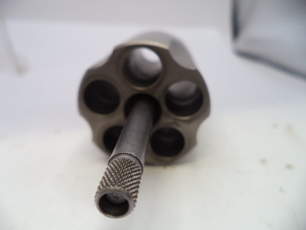 A close-up shot of a metallic revolver cylinder and barrel, showing the six internal chambers and a knurled barrel tip, resting on a white surface.