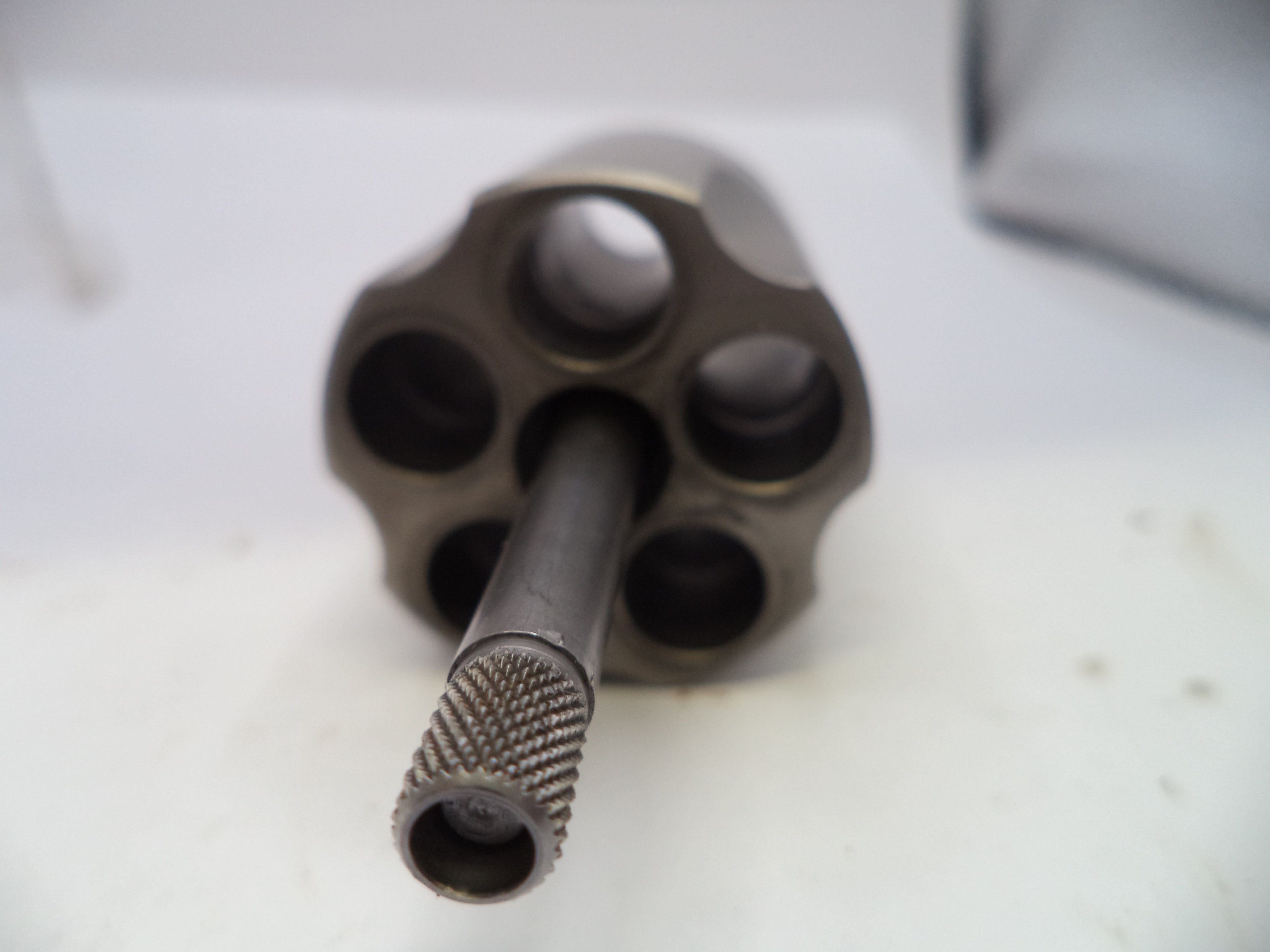 A close-up shot of a metallic revolver cylinder and barrel, showing the six internal chambers and a knurled barrel tip, resting on a white surface.