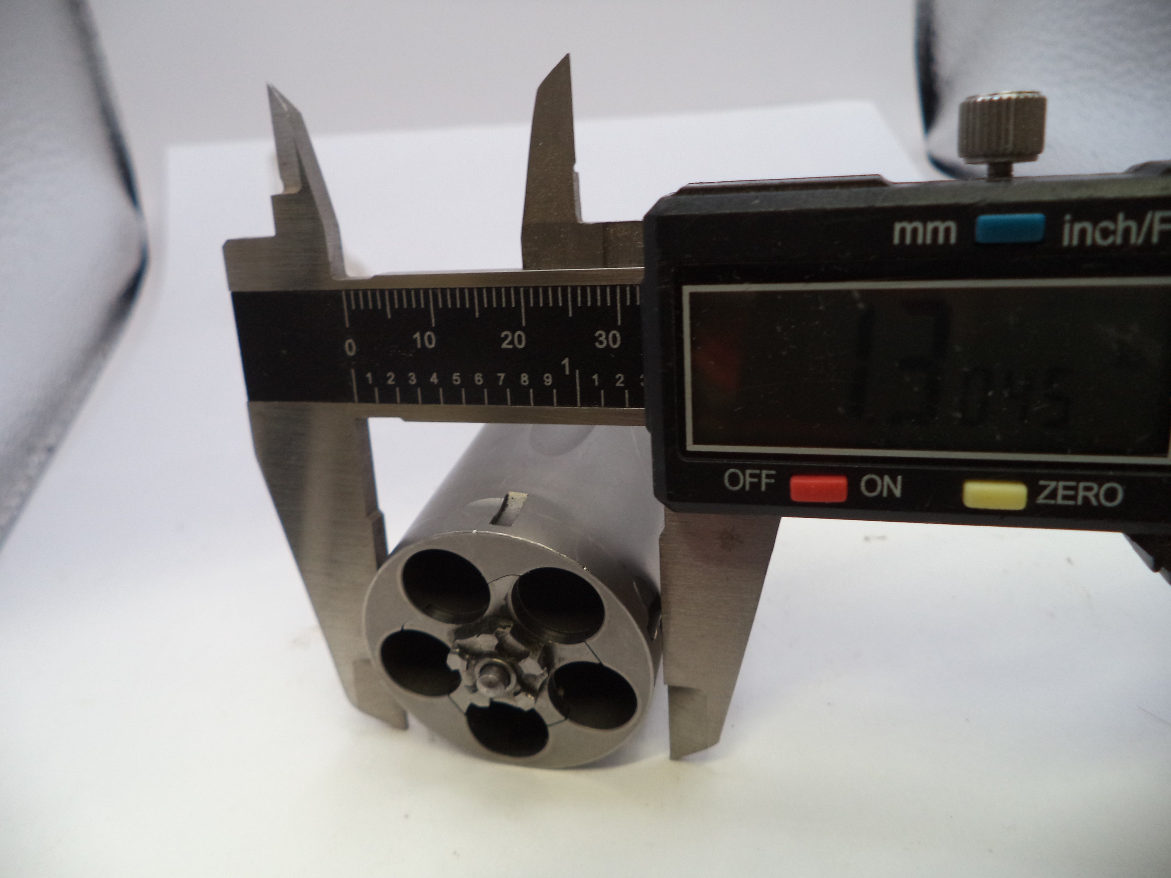 A close-up of a digital caliper measuring the diameter of a small cylindrical metal component with five circular openings.
