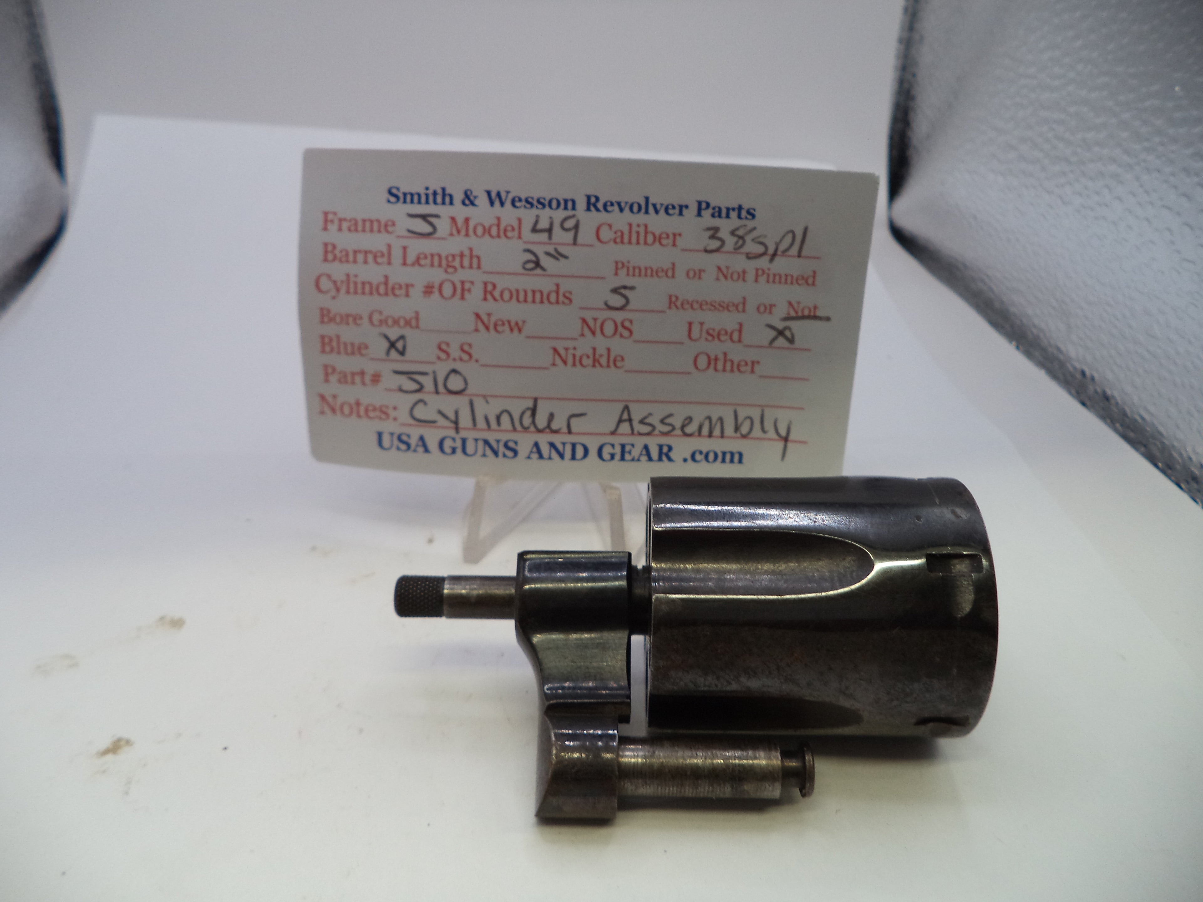 A used metal cylinder assembly for a Smith & Wesson revolver, displayed on a white surface with a paper parts card in the background.