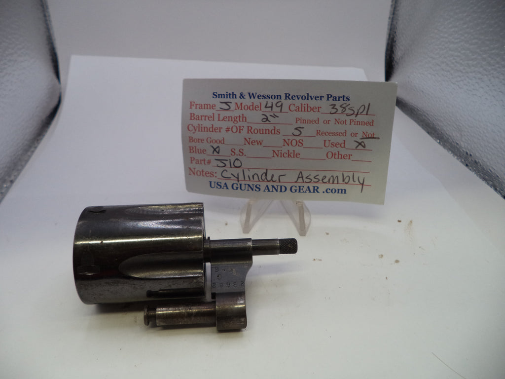 A used metal cylinder assembly for a Smith & Wesson revolver, displayed on a white surface with a handwritten identification card behind it.