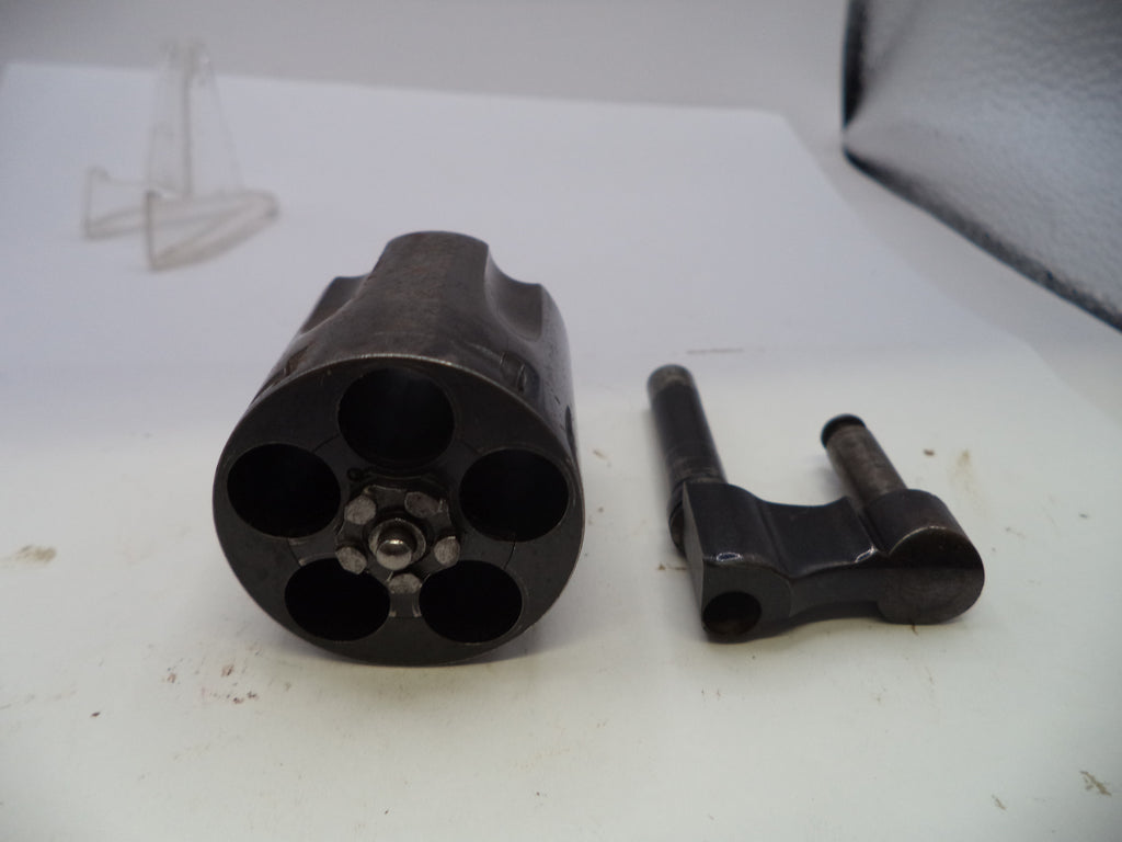 A dark metallic revolver cylinder disassembled to show the six internal chambers, alongside a corresponding black metal firing pin block, photographed on a white surface.