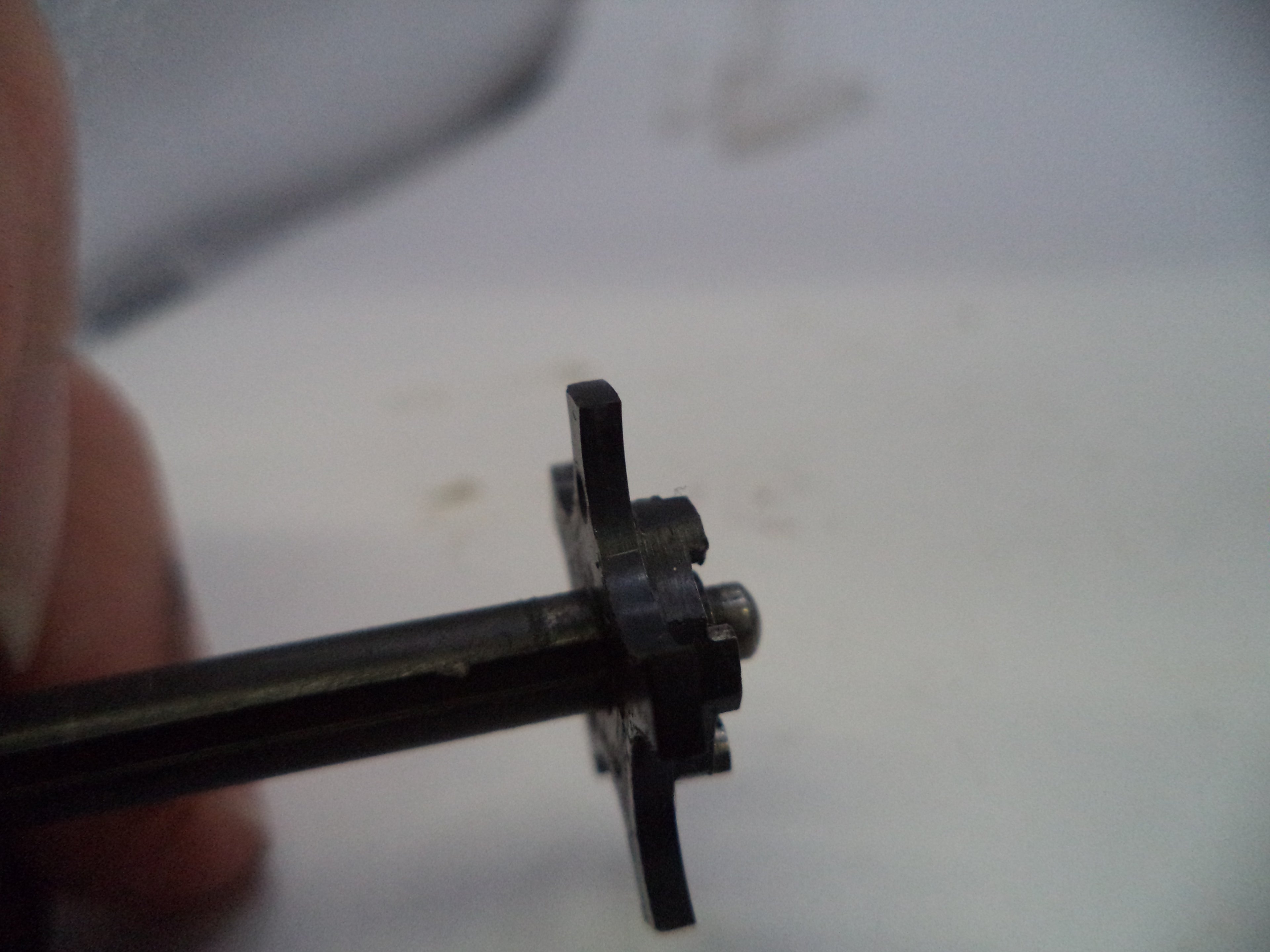 A close-up of a small black metal mechanical component held by fingers against a light, out-of-focus background.