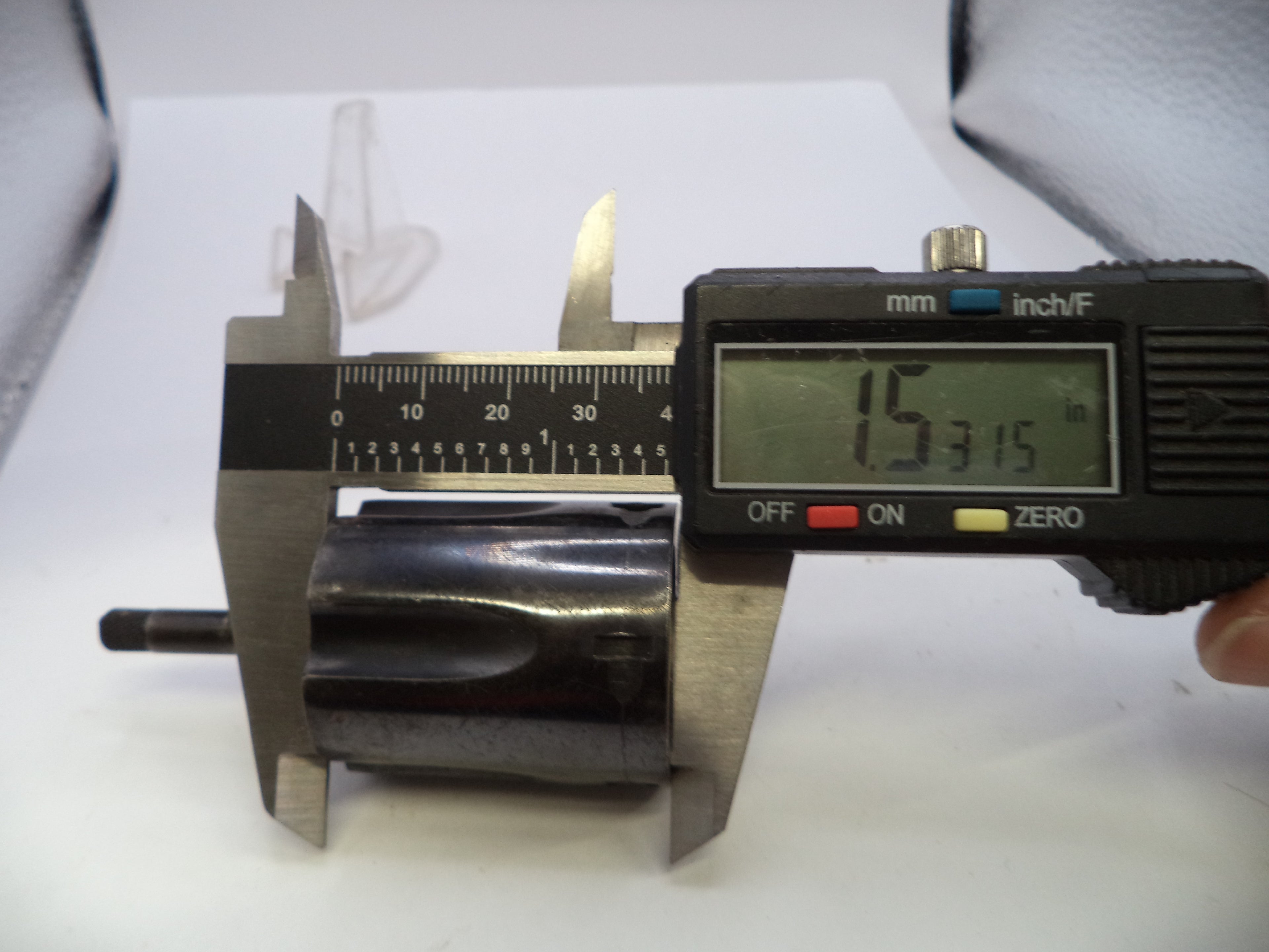 A digital caliper measuring a dark metallic cylindrical component on a white surface, with a hand holding the caliper's black display unit.