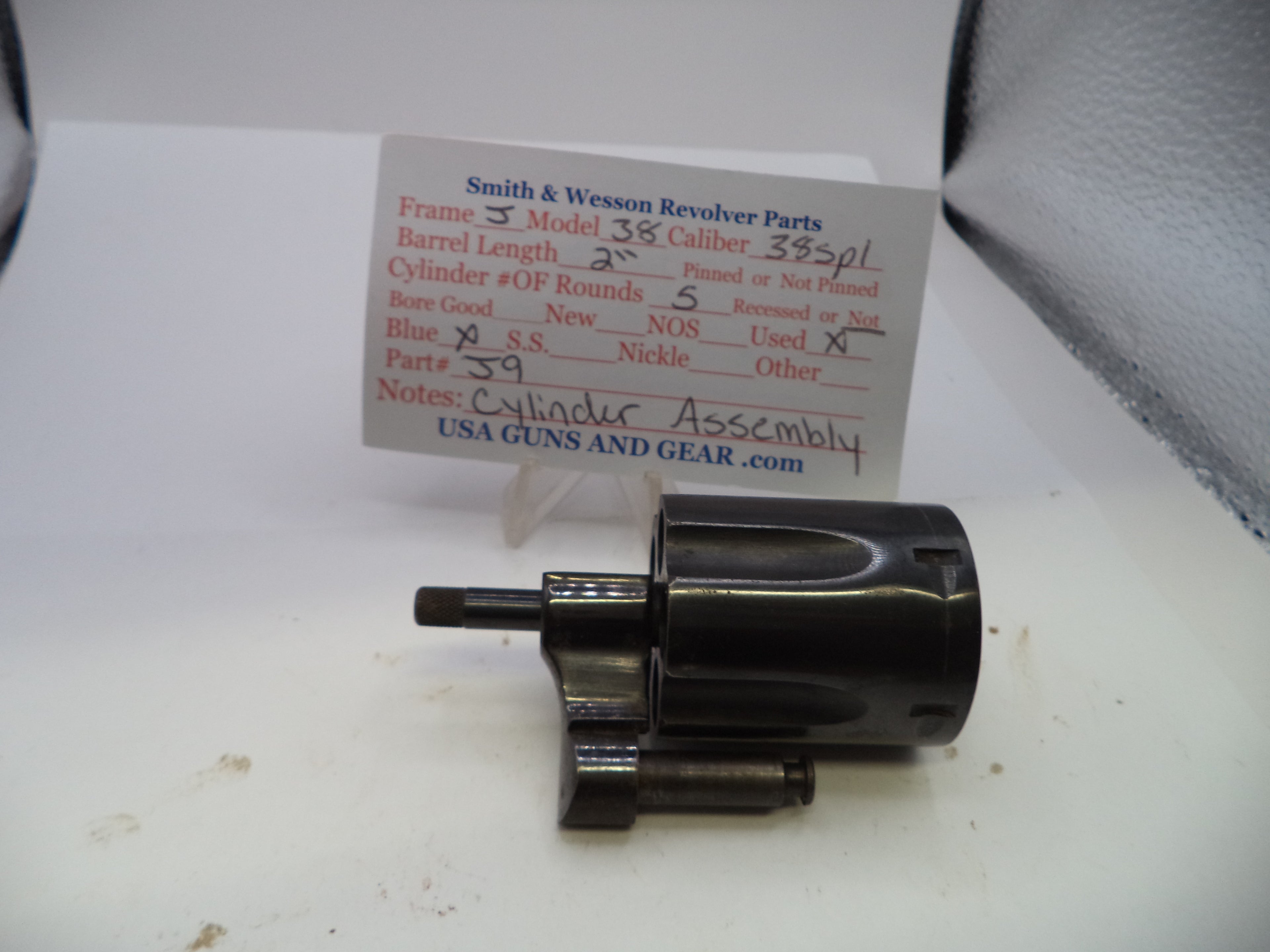 A black metal revolver cylinder assembly part, photographed on a white surface with a handwritten parts label card behind it reading Smith & Wesson Revolver Parts.