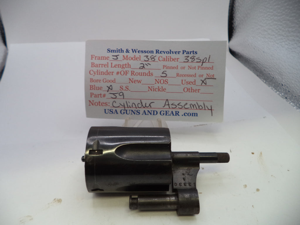 A used black metal Smith & Wesson revolver cylinder assembly part, shown in front of a white paper label card with handwritten specifications.