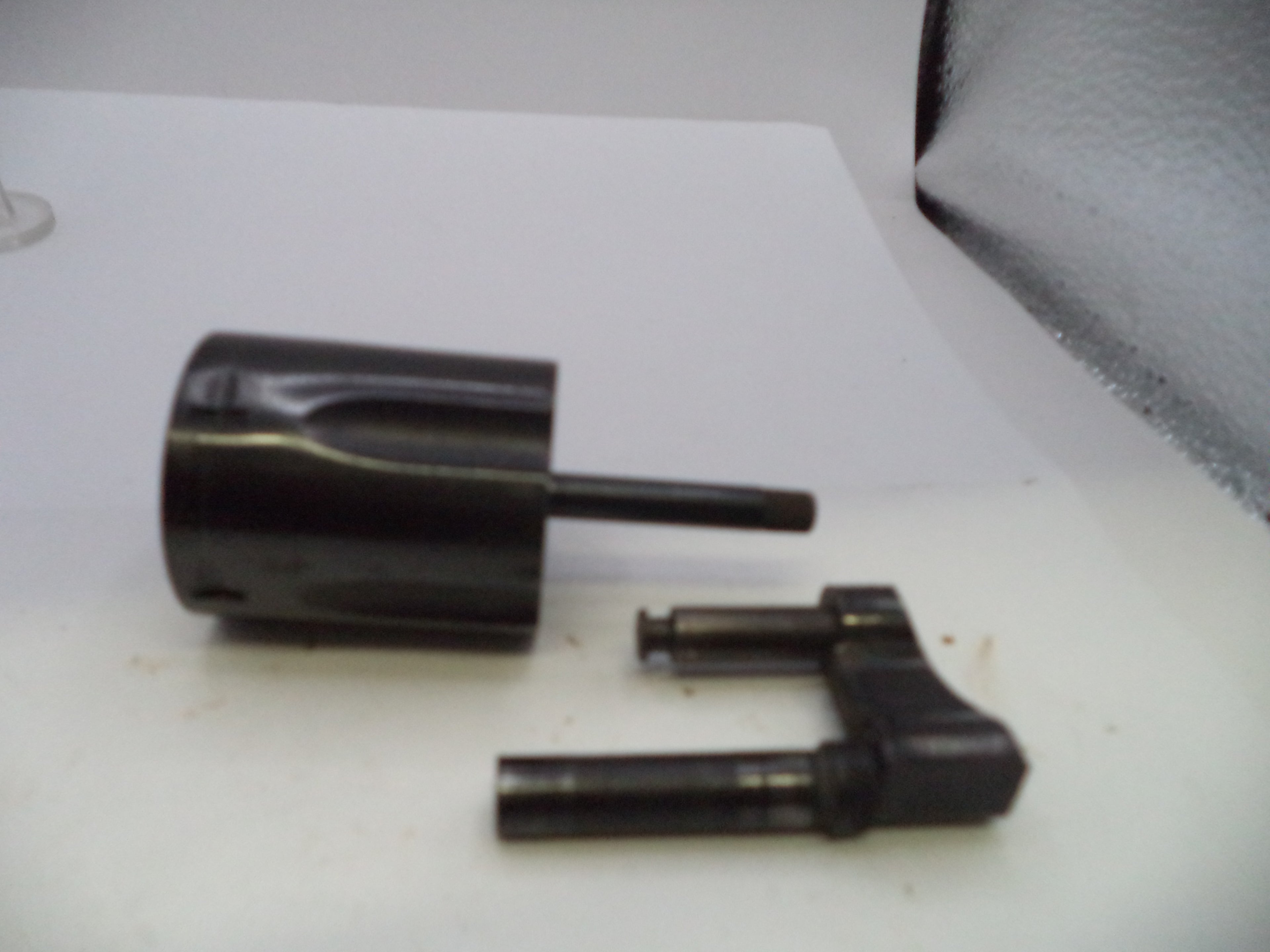 A black plastic or metal cylindrical component with a protruding rod, shown alongside a separate matching black mechanical piece on a white surface.