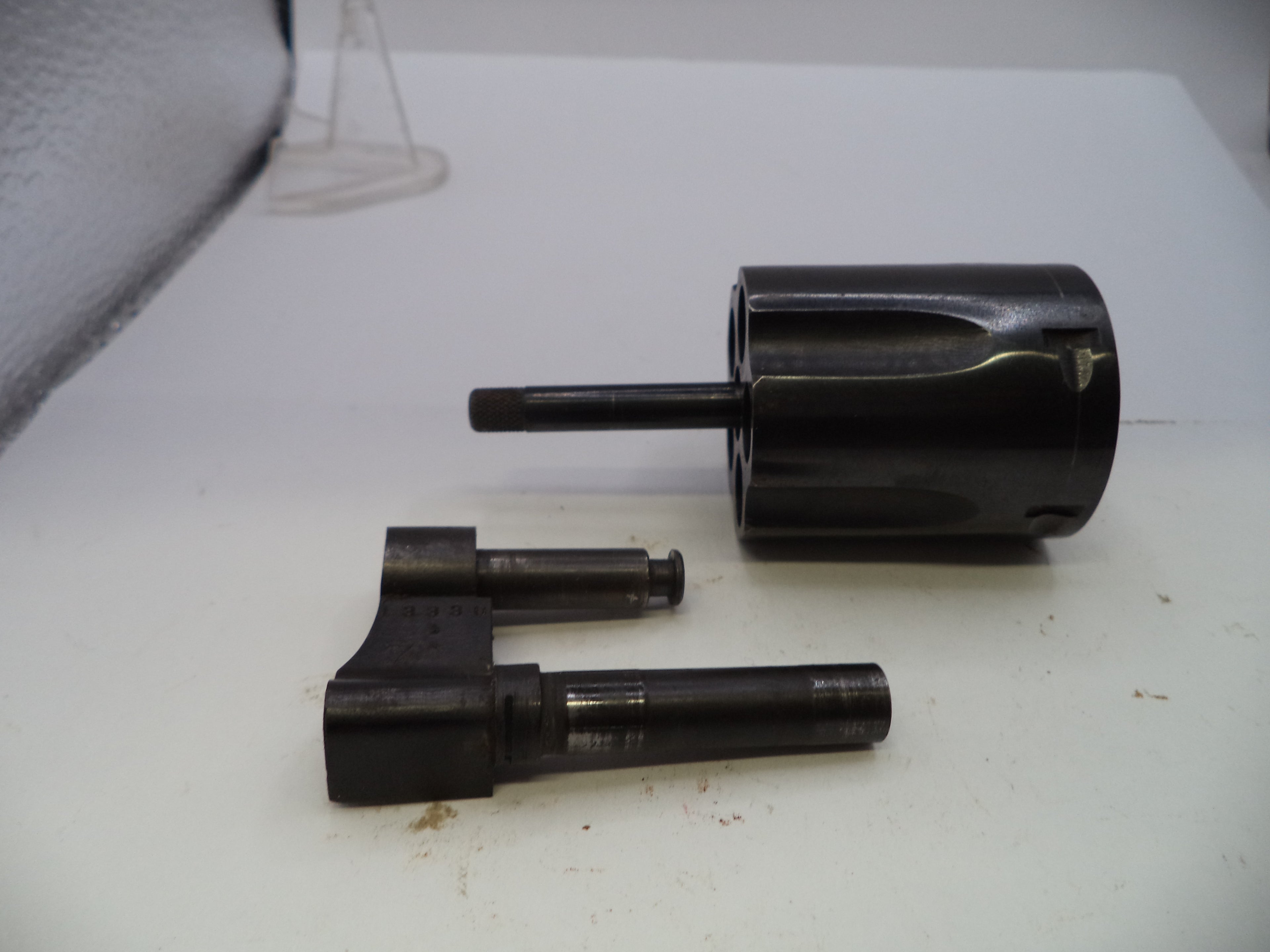 A set of dark metallic mechanical components, including a cylindrical sleeve with a protruding shaft and a matching bracketed rod assembly, photographed on a white surface.