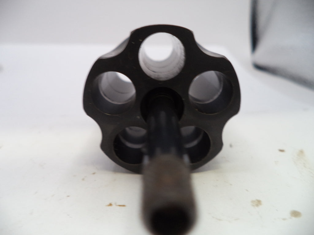 A close-up shot of a black metal revolver cylinder with six circular openings and a central barrel shank, resting on a white surface.