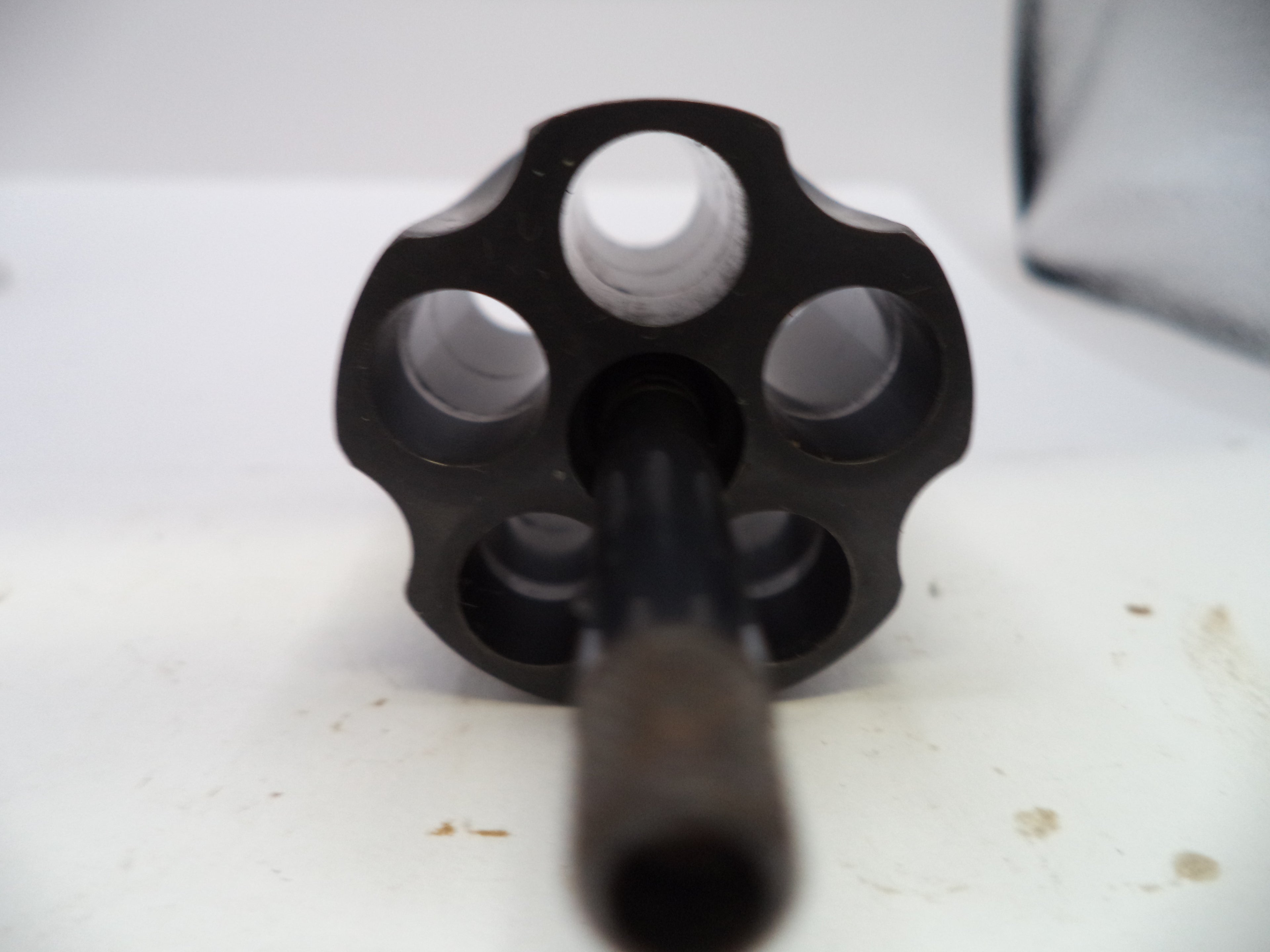 A close-up shot of a black metal revolver cylinder with six circular openings and a central barrel shank, resting on a white surface.