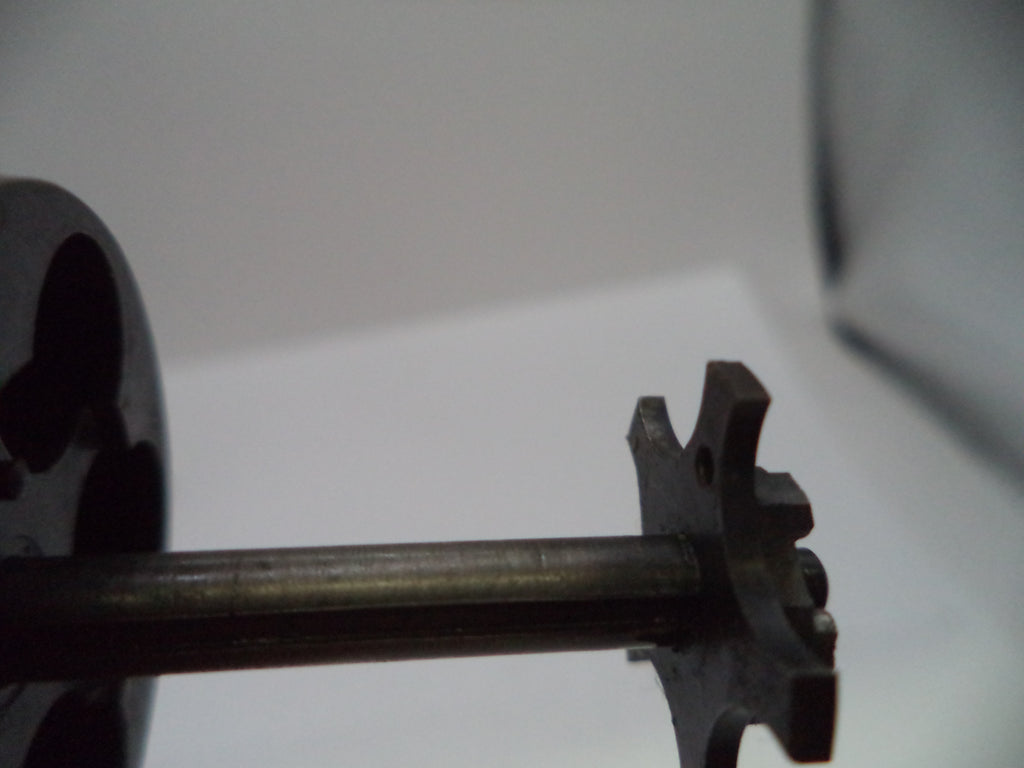 A close-up of a dark metal gear or sprocket attached to a horizontal shaft, photographed against a plain light background.