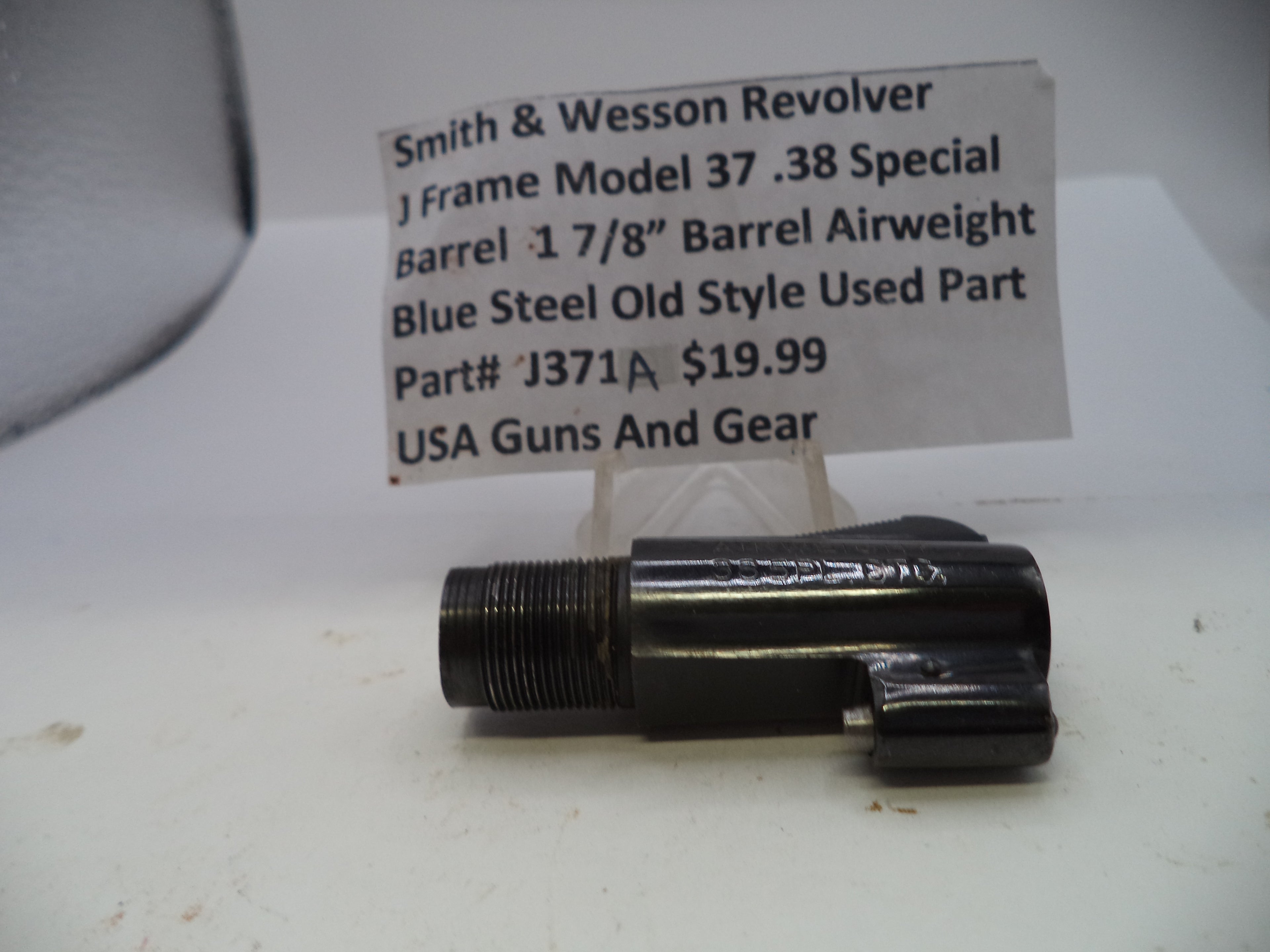 A black metal Smith & Wesson revolver barrel airweight part, shown on a white surface with a paper label card in the background.