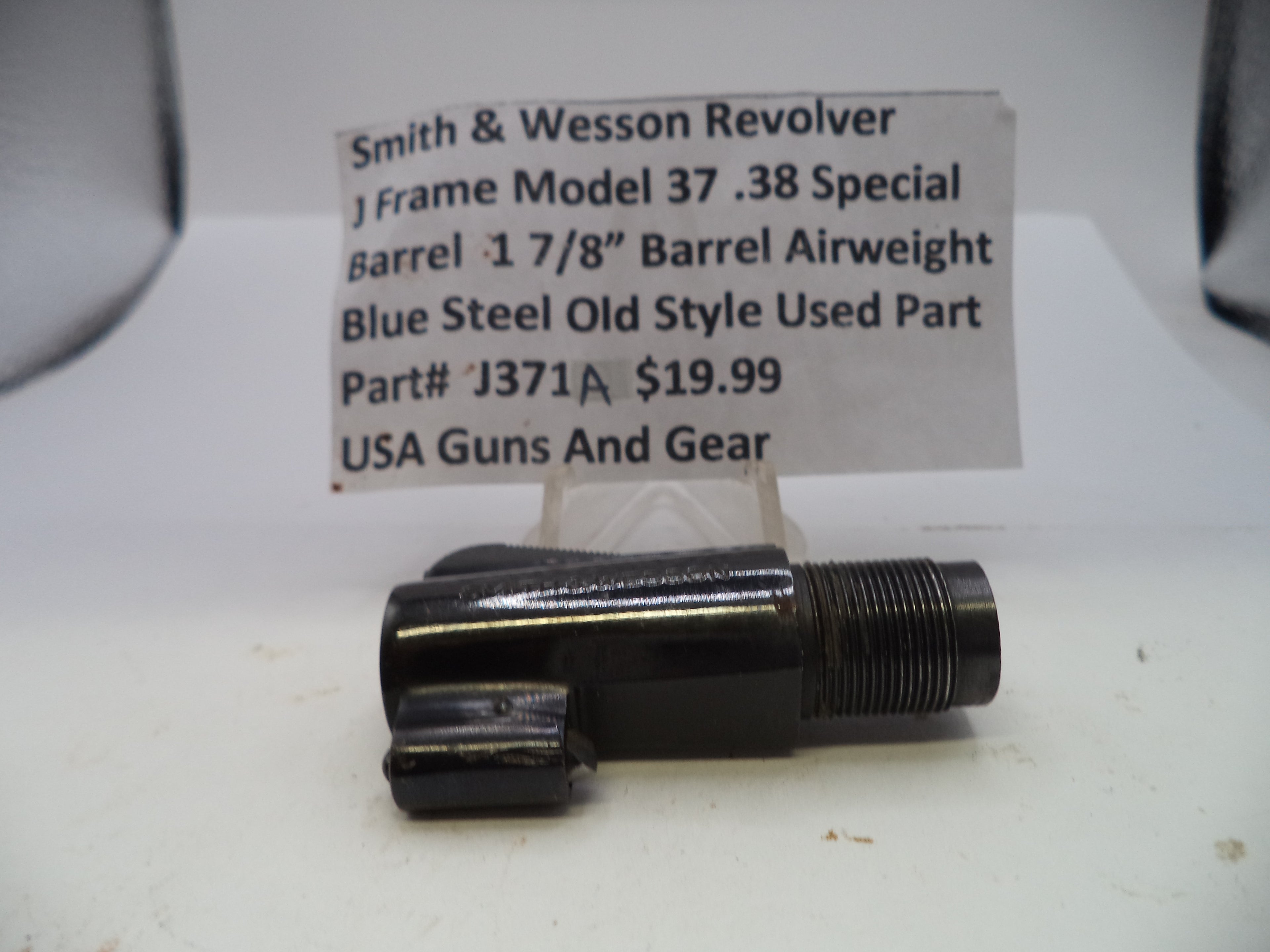 A used black metal Smith & Wesson revolver barrel part with a threaded end, displayed on a white surface with a handwritten price tag card behind it.