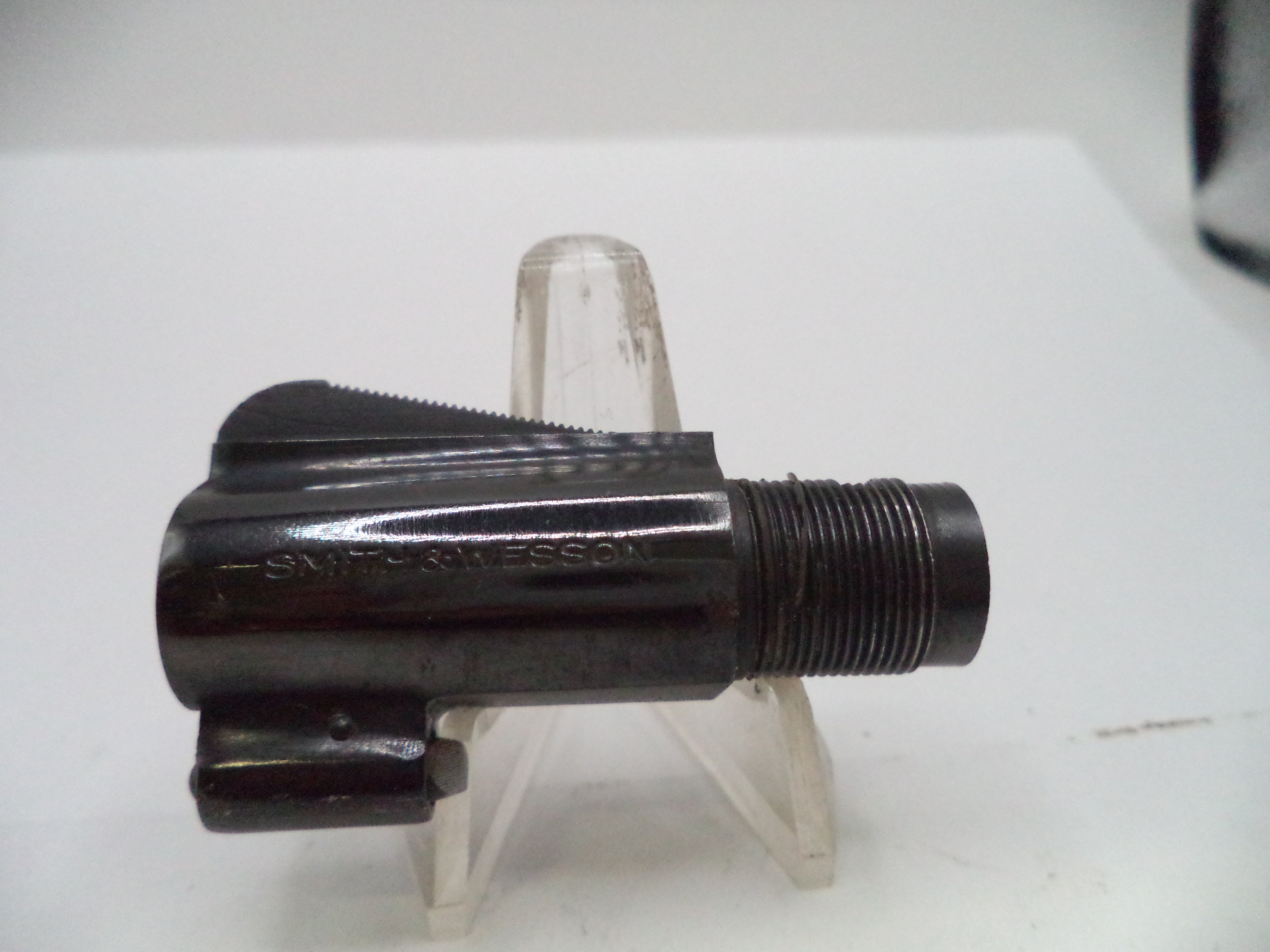 A black metal threaded component, likely a firearm magazine or suppressor part, resting on a clear plastic display stand against a white background.