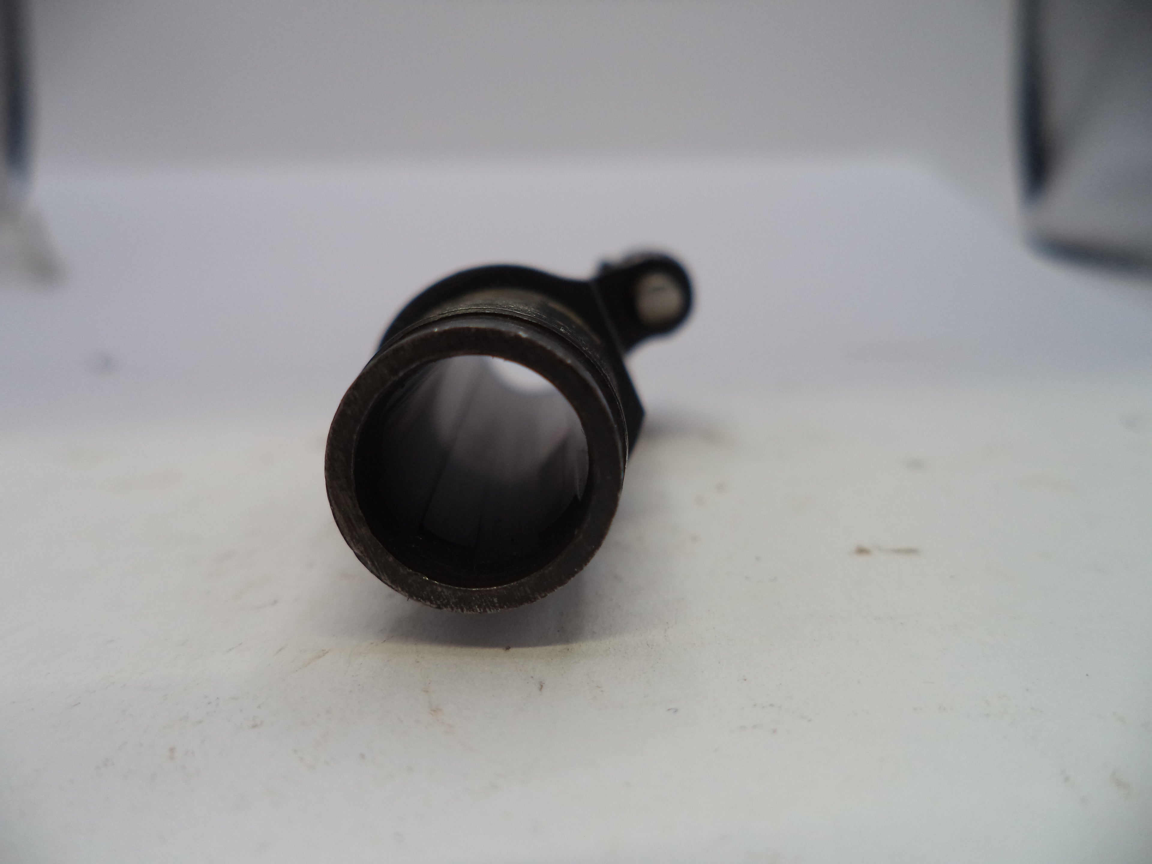 A close-up of a dark metal cylindrical component with a circular opening and a small protruding tab, resting on a white surface.