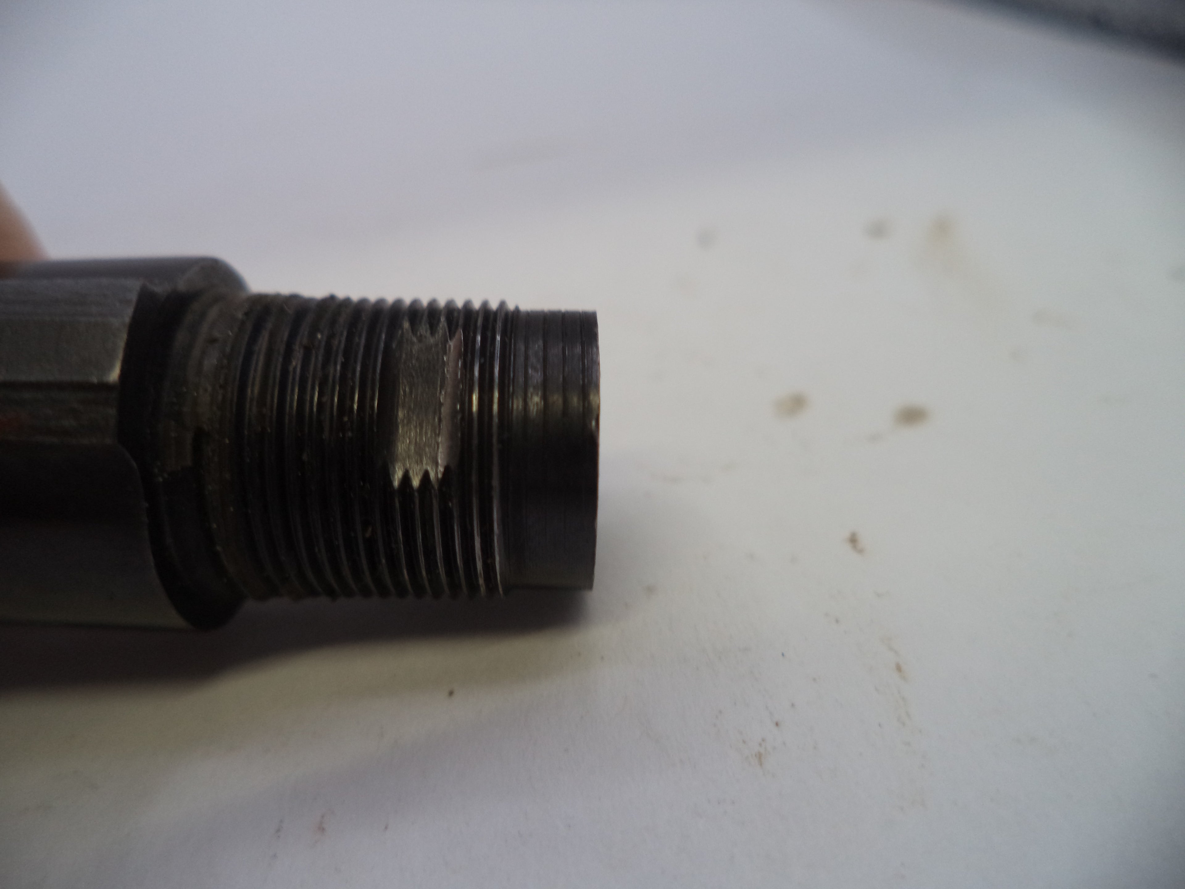 A close-up of a dark metal threaded component, possibly a mechanical fitting or valve stem, resting on a white surface.