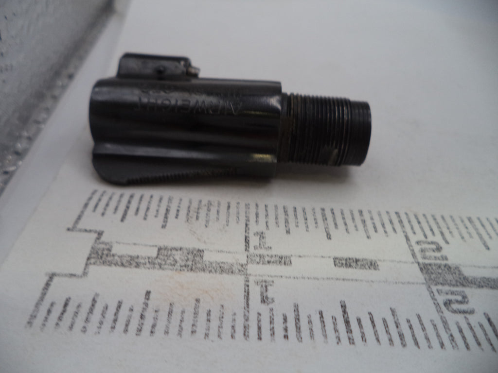 A small black metal threaded component with a small attached rectangular piece, photographed on a white surface with a printed ruler or measurement guide underneath for scale.