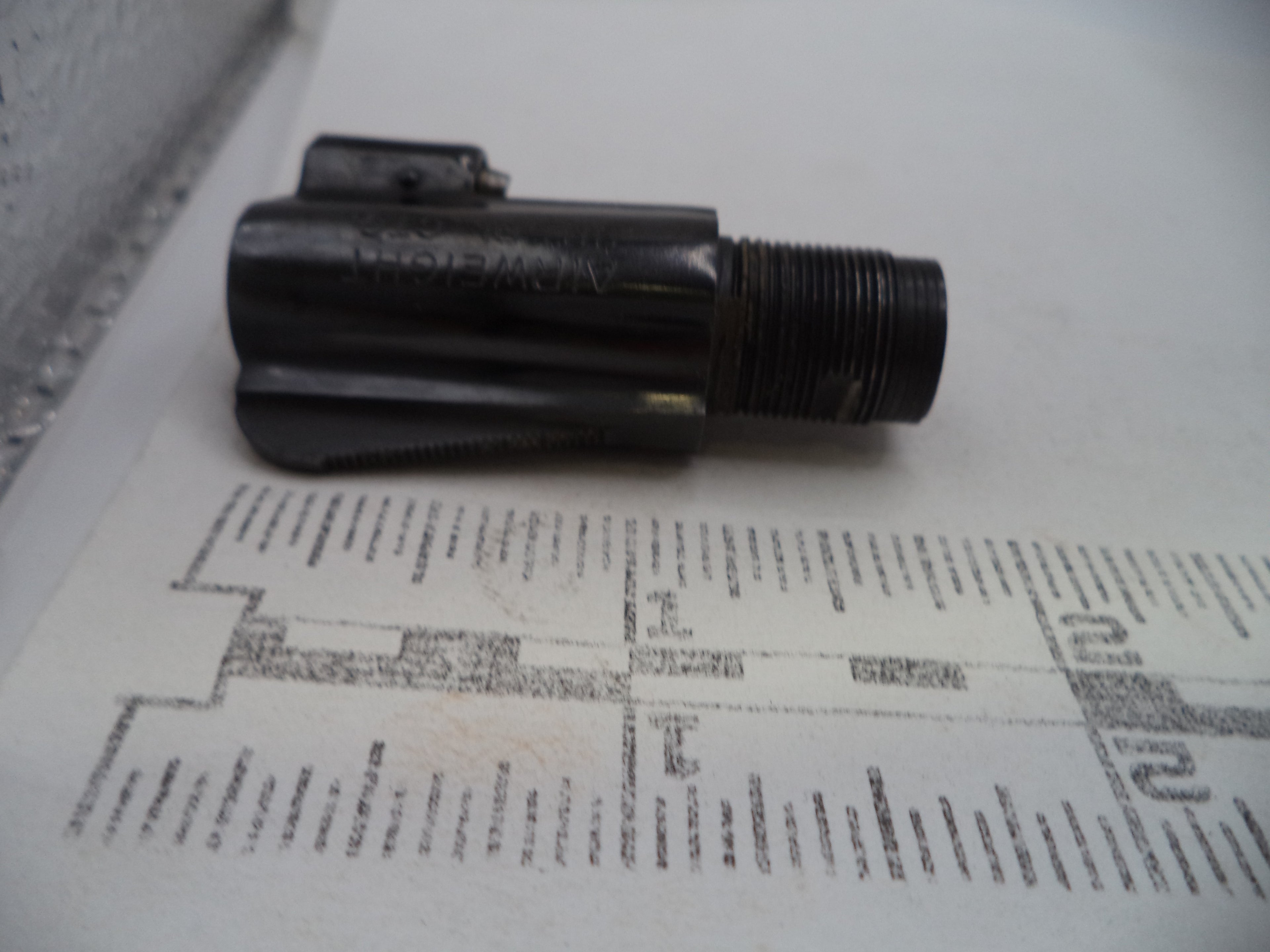 A small black metal threaded component with a small attached rectangular piece, photographed on a white surface with a printed ruler or measurement guide underneath for scale.