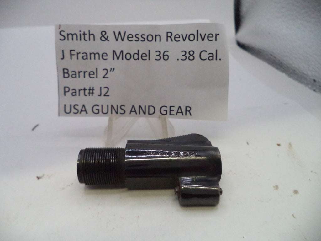 A black metal Smith & Wesson revolver barrel adapter part, model J2, shown on a white surface with a handwritten label card in the background.