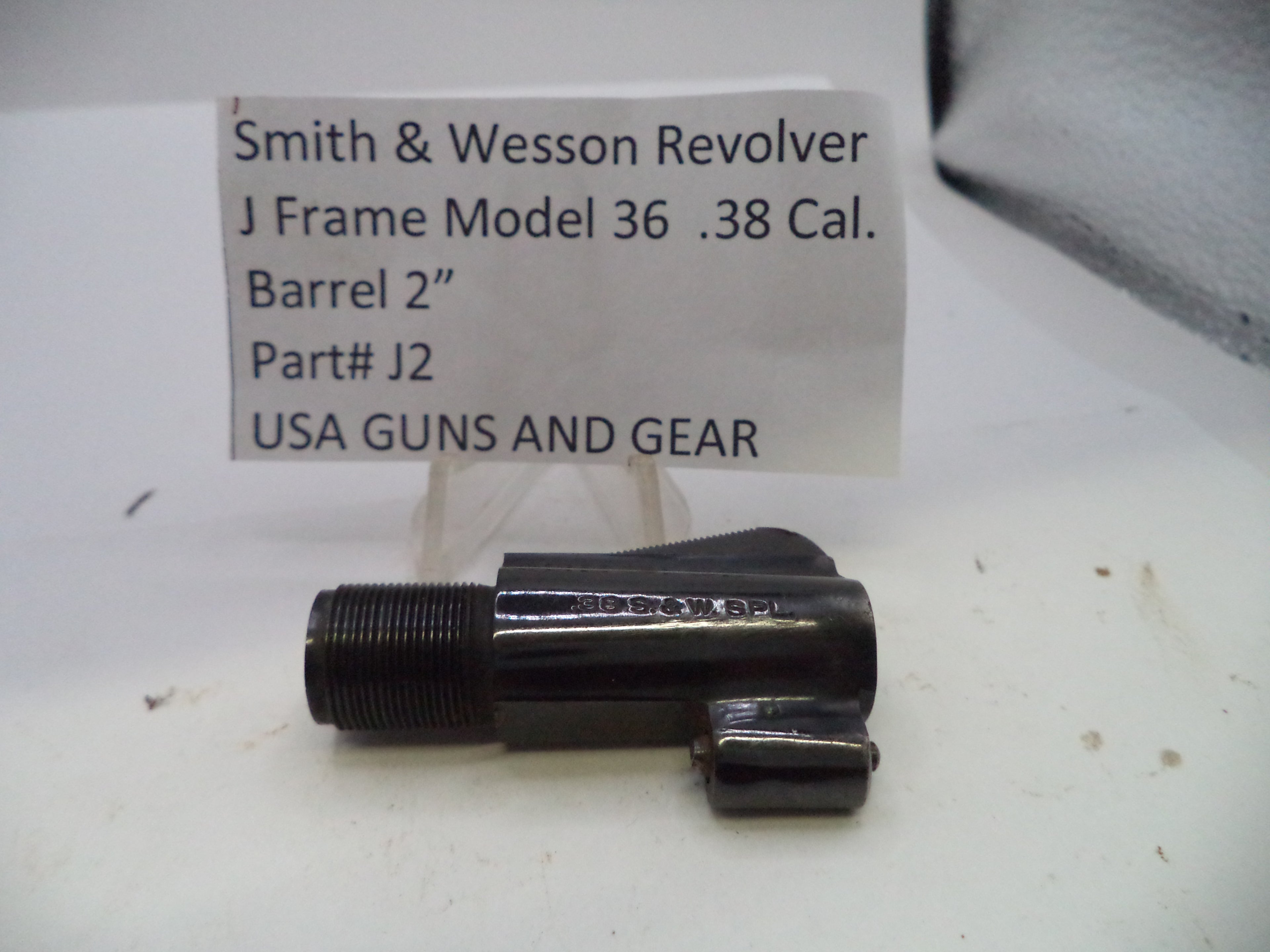 A black metal Smith & Wesson revolver barrel adapter part, model J2, shown on a white surface with a handwritten label card in the background.